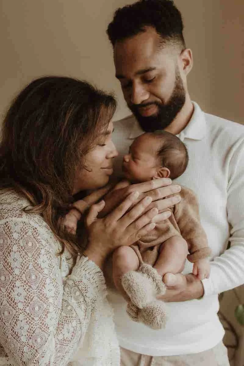 A family of three, a mother, father, and a newborn, sharing a tender moment indoors with the mother and father lovingly holding their baby.
