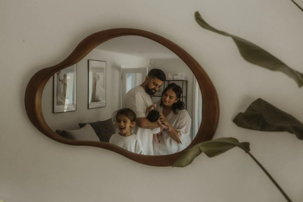 A family of four looking at a newborn baby through a mirror, with framed art on the wall behind them.