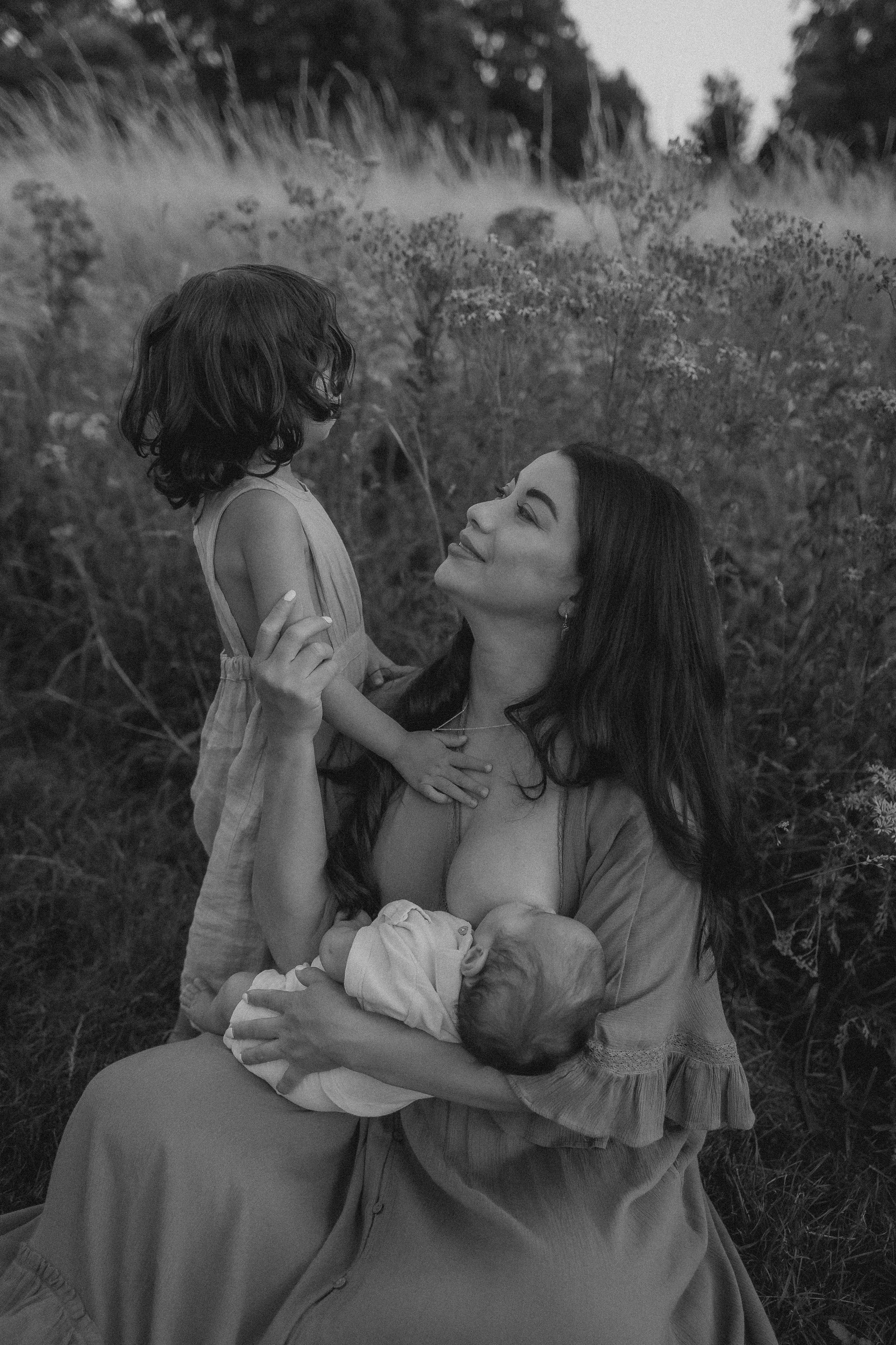 A woman with long hair holding a baby in her arms, while a young girl stands in front of her outside among bushes and trees, smiling and looking at each other.