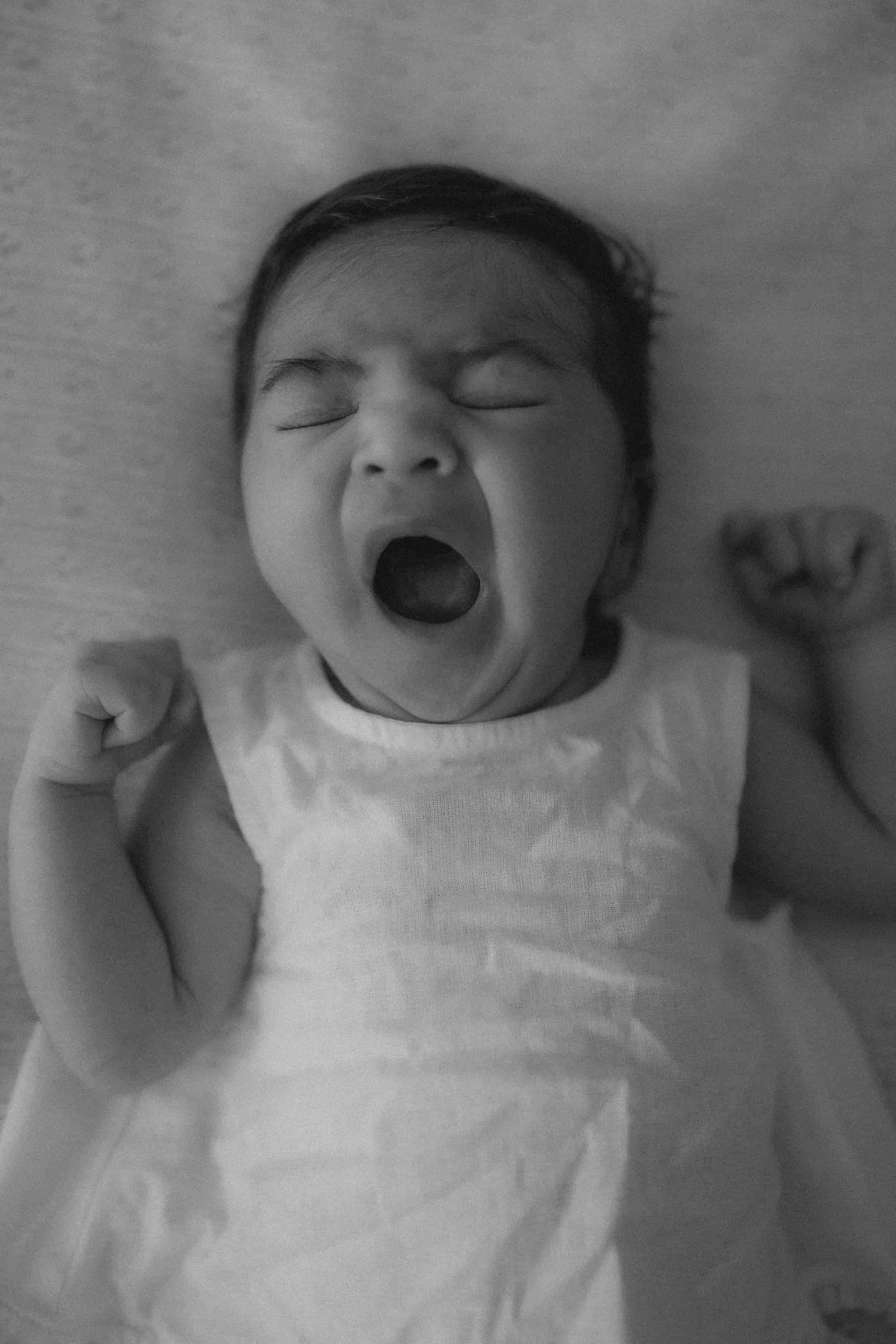 A black and white photo of a sleeping baby lying on a soft surface, with fists clenched near the shoulders, mouth open, and eyes closed.