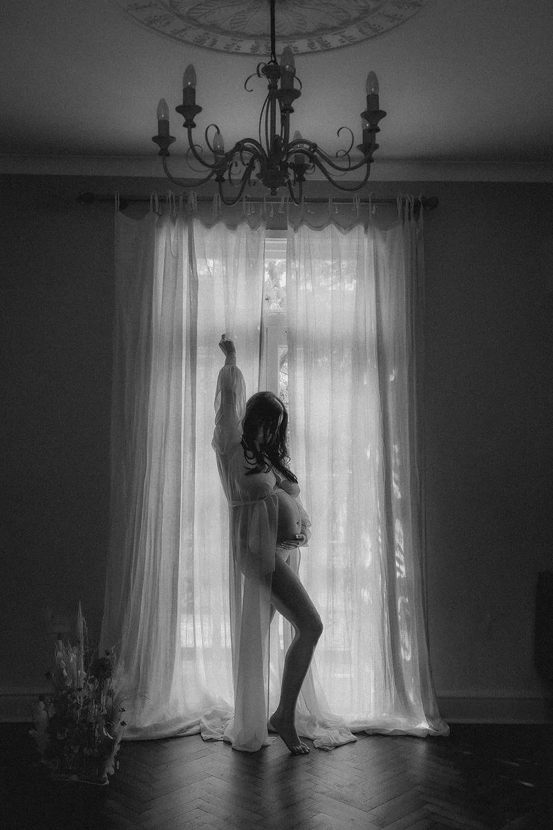 A pregnant woman standing by a window with sheer curtains, with her arm raised, in a softly lit room with a chandelier hanging from the ceiling.