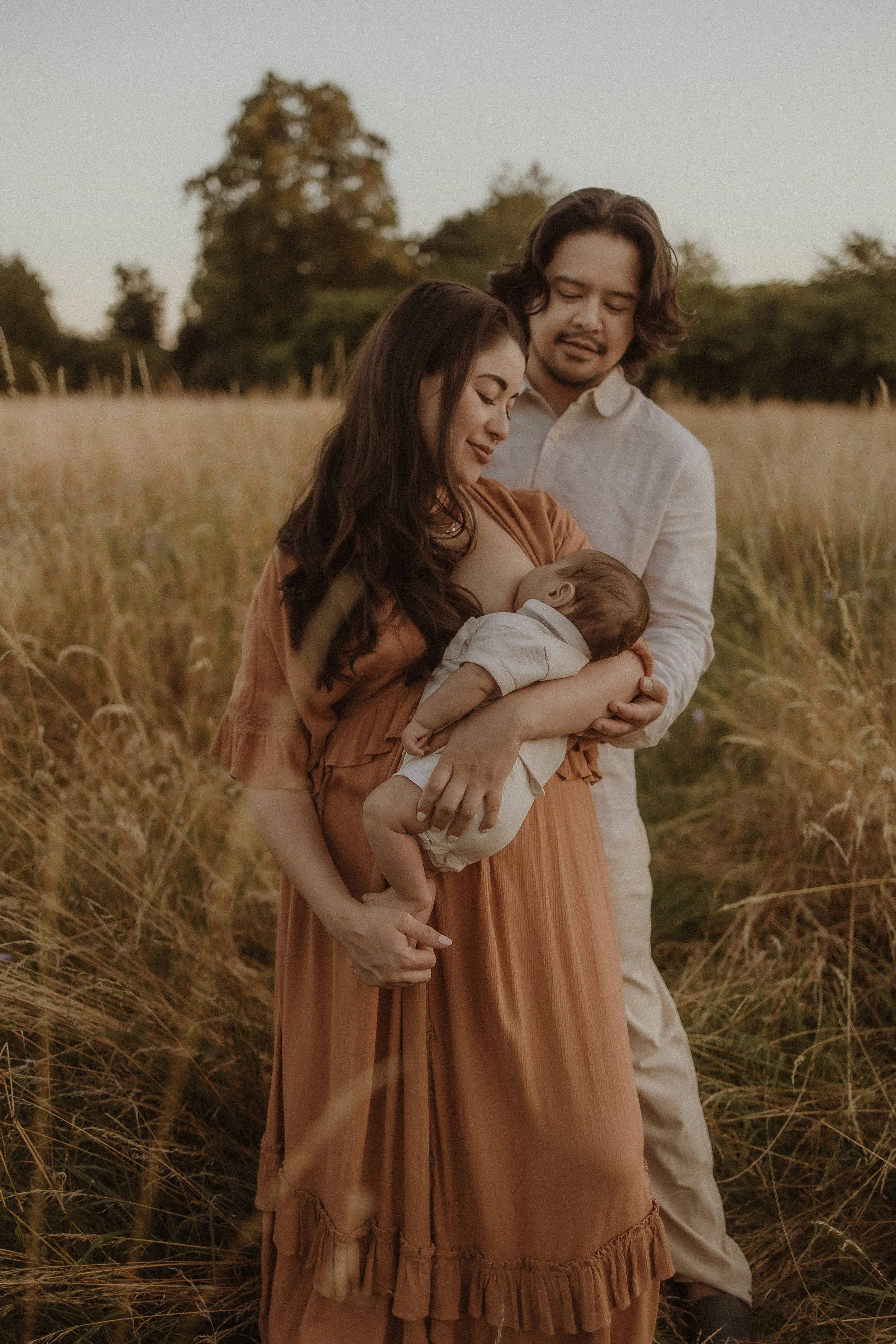 A family of three standing in a field of tall grass, with a woman holding a sleeping baby and a man standing behind her.