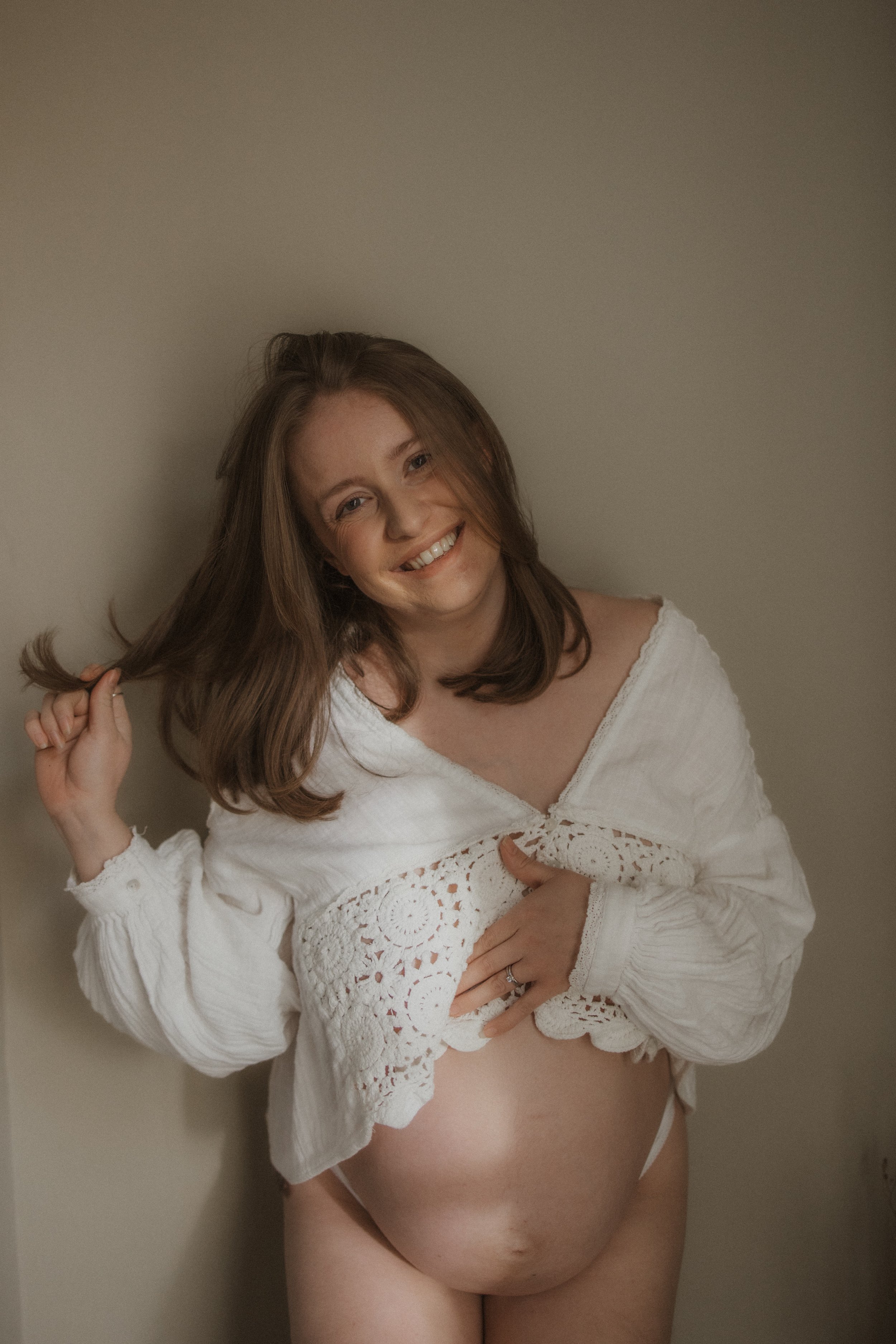 A pregnant woman with long brown hair, smiling, lifting her white lace-sleeve shirt to reveal her baby bump, standing against a plain wall.