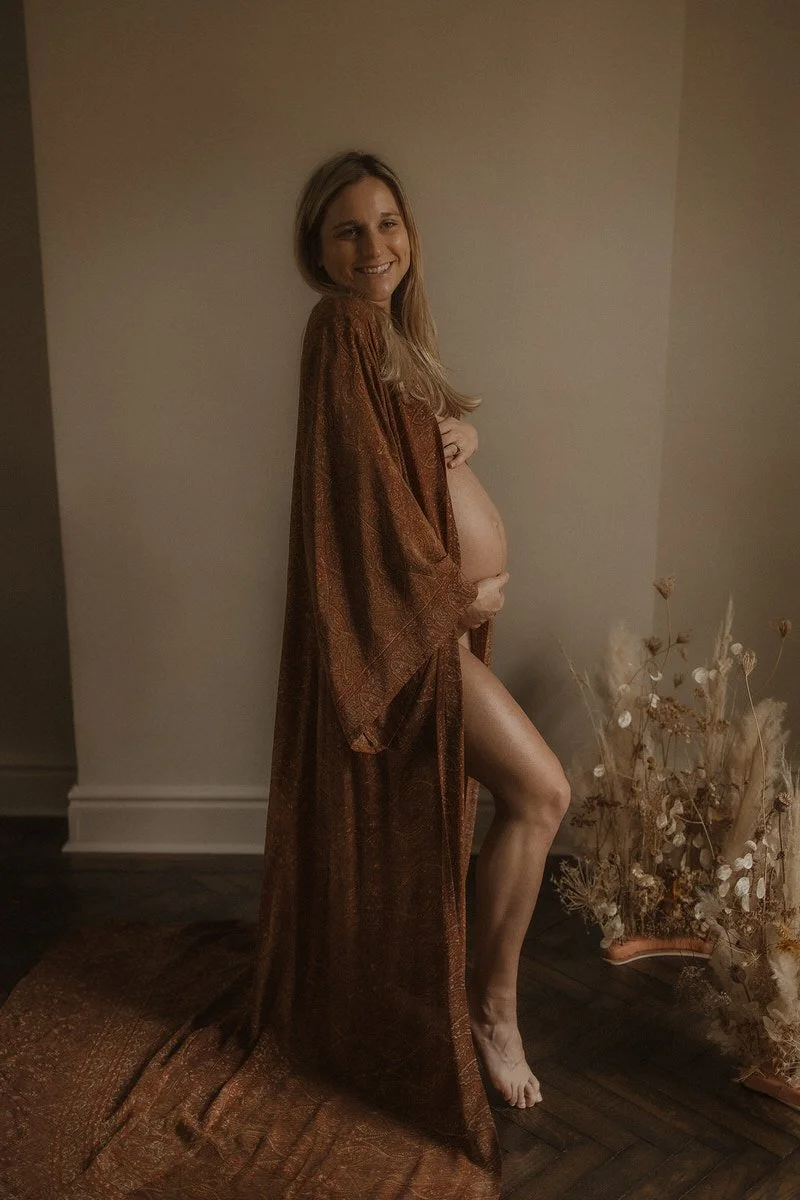 A pregnant woman standing indoors, wearing a long brown patterned robe, smiling and holding her belly, with a vase of dried flowers on the floor nearby.