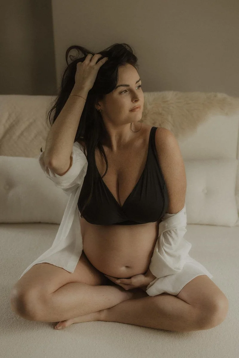 A pregnant woman sitting cross-legged on a bed, looking to the side, with her hand in her dark hair, wearing a black bra and a white shirt open at the front.