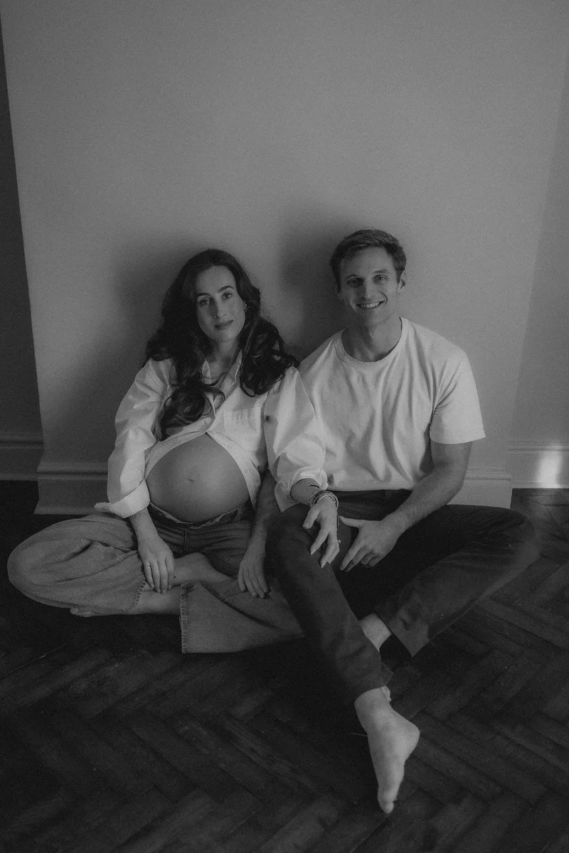 A pregnant woman and a man sitting on the floor against a plain wall, smiling.