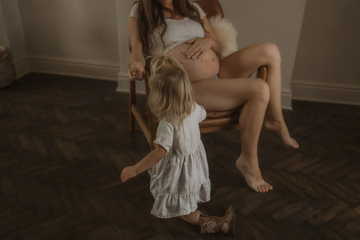 A pregnant woman sitting in a chair with her hand on her belly, surrounded by colorful stickers, while a young girl, wearing a white dress and brown cowboy boots, stands in front of her, touching her belly.