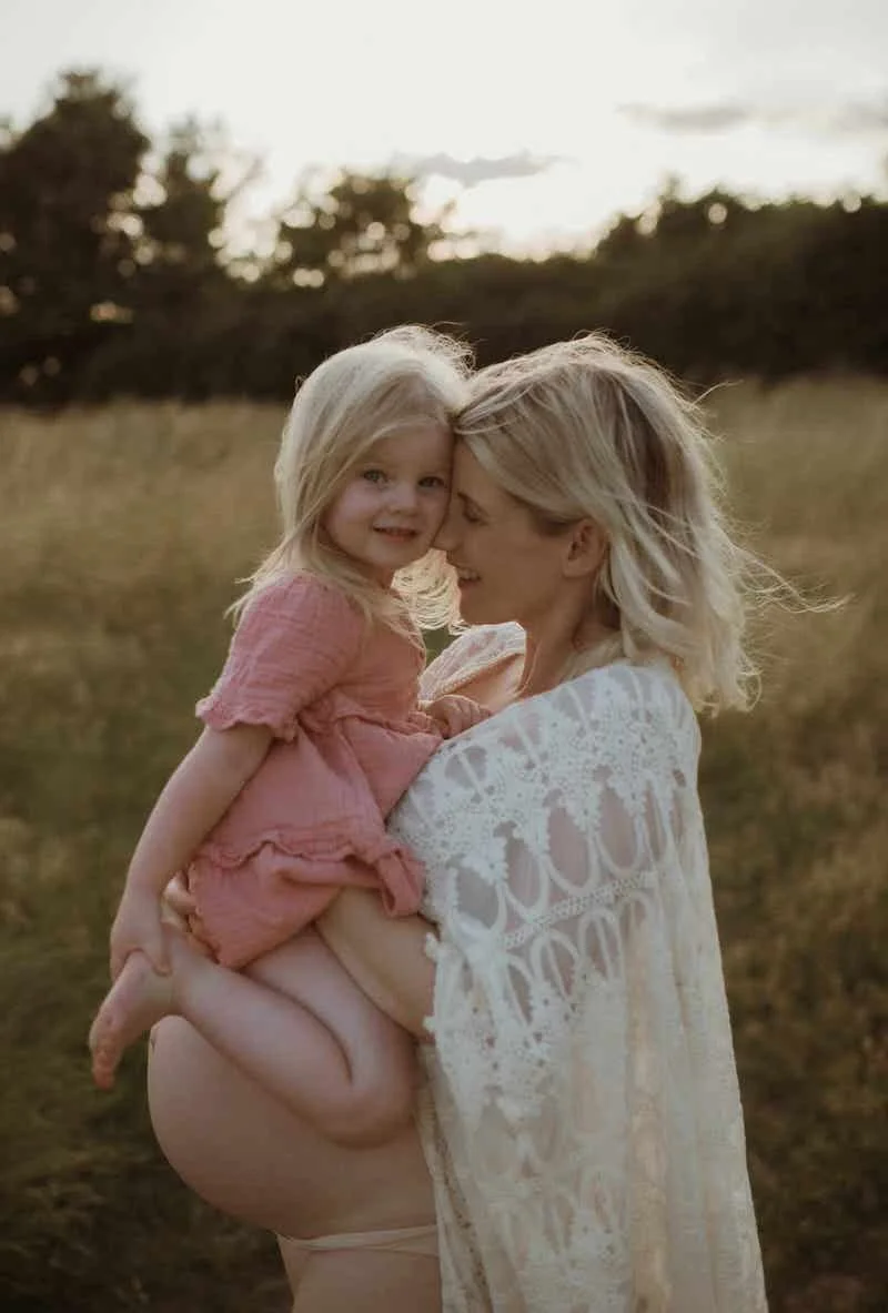 A woman holding a young girl in an outdoor field at sunset; they are touching foreheads and smiling.