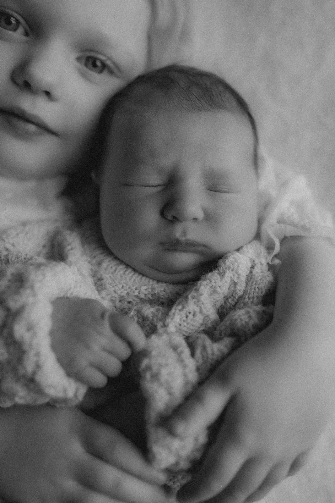 A young child and a newborn baby, both with light skin, lying close together. The older child looks directly at the camera with a gentle expression, while the baby is peacefully sleeping, wrapped in a knitted blanket.