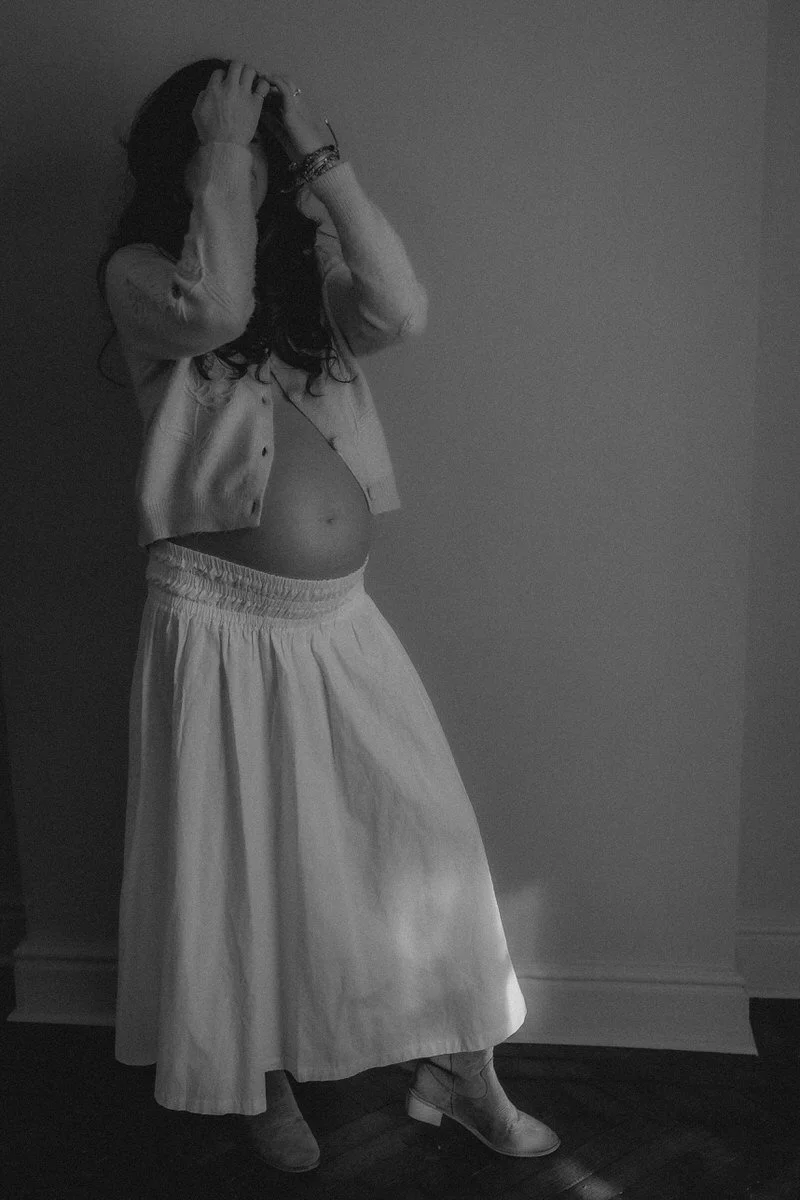 A woman with long dark hair standing against a plain wall, wearing a light-colored blouse, high-waisted skirt, and heeled shoes, with her hands in her hair.