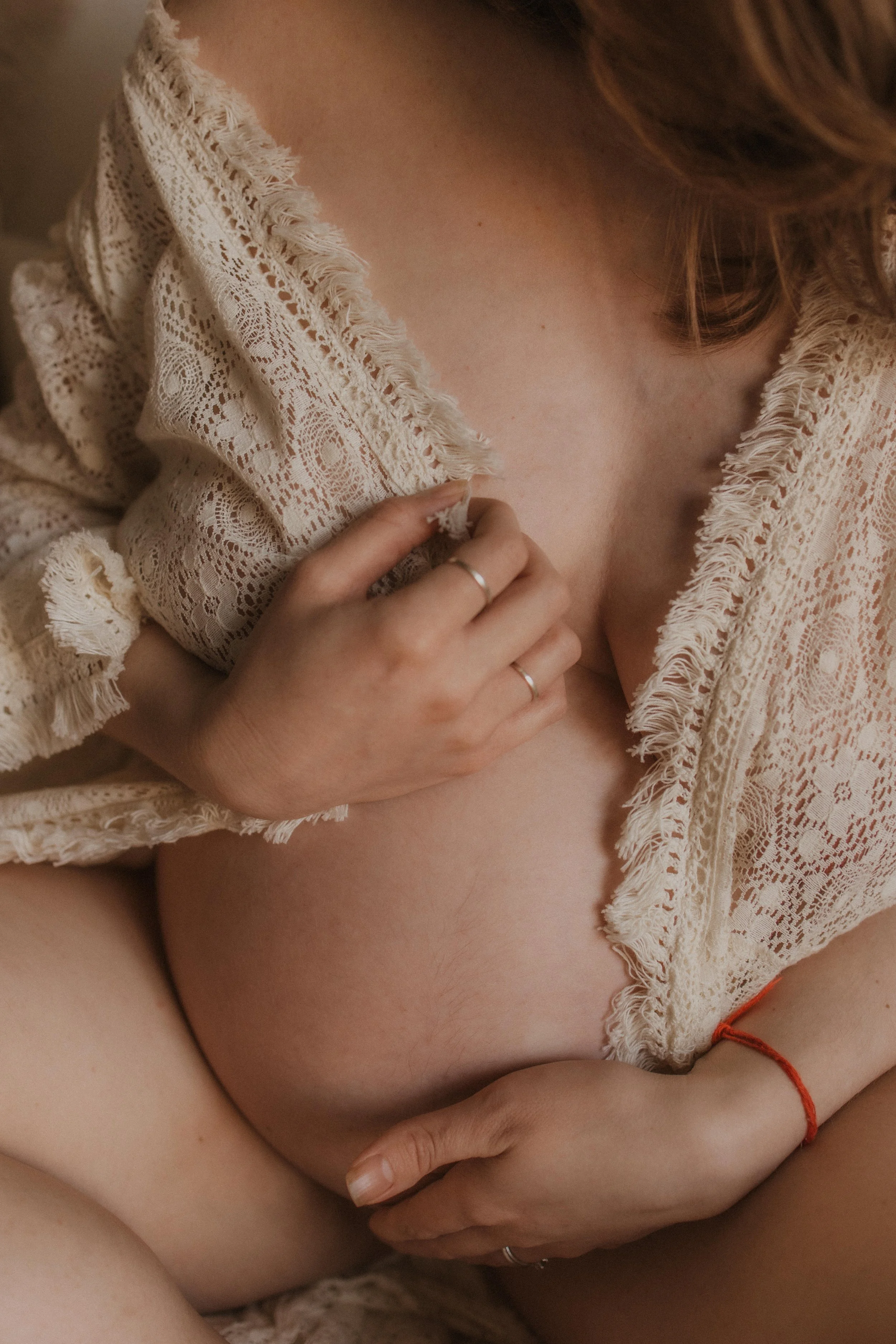 A woman wearing a lacy beige top, holding her pregnant belly with one hand and gently touching her chest with the other. The focus is on her hands and abdomen.