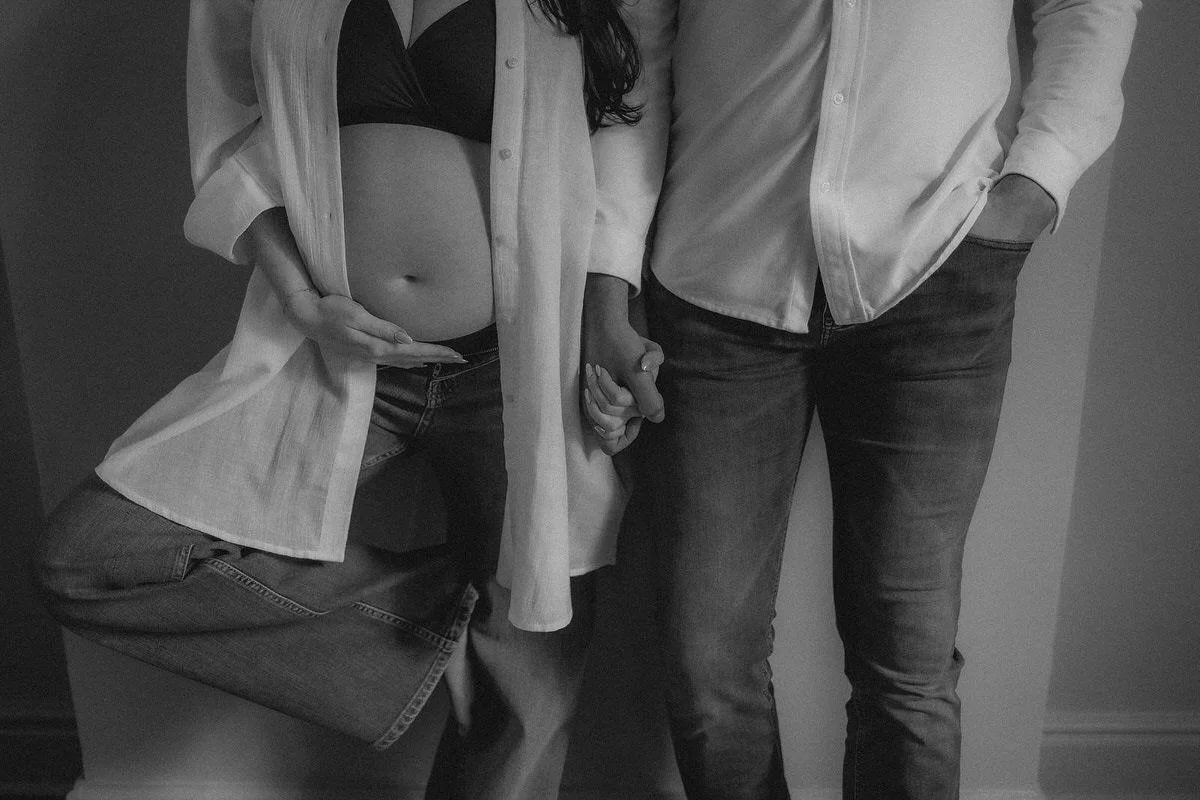 A pregnant woman and a man holding hands, with only their torsos and legs visible, standing next to each other in a room.