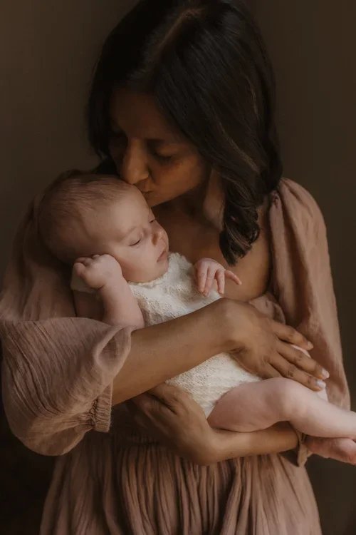 Woman holding a sleeping baby close, gently kissing the baby's forehead in a warm, intimate moment.