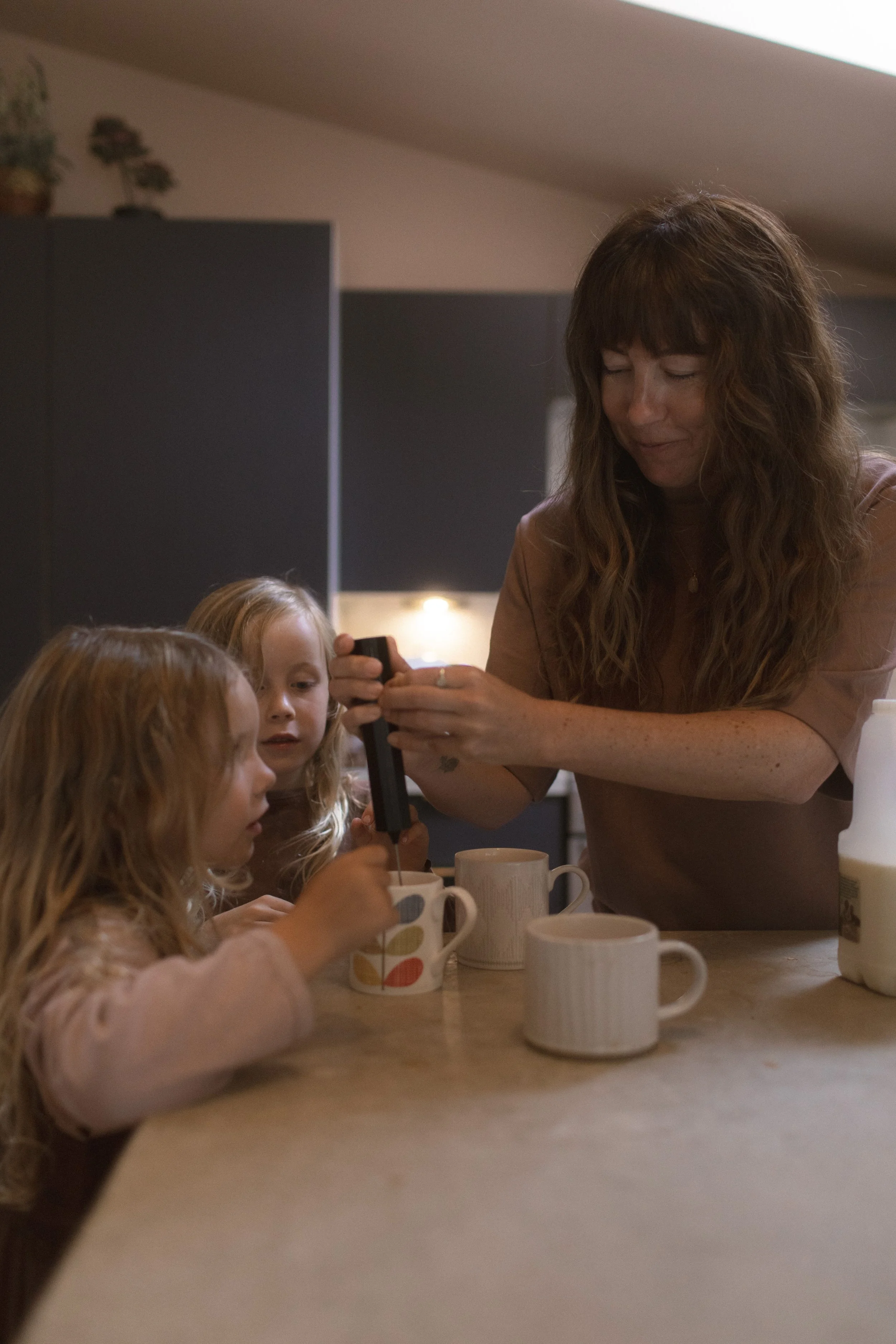 A woman and two young girls at a kitchen counter, with the woman pouring a beverage into mugs.