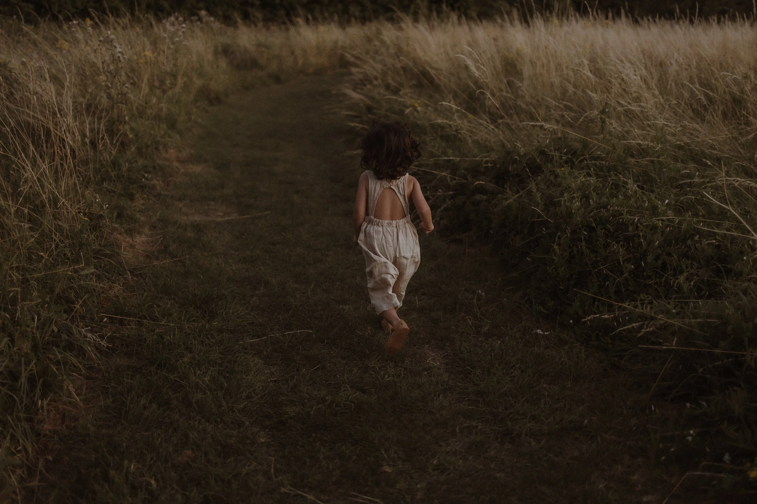 A young girl with curly hair running on a narrow grassy trail through tall dry grass in a field during sunset or early evening.