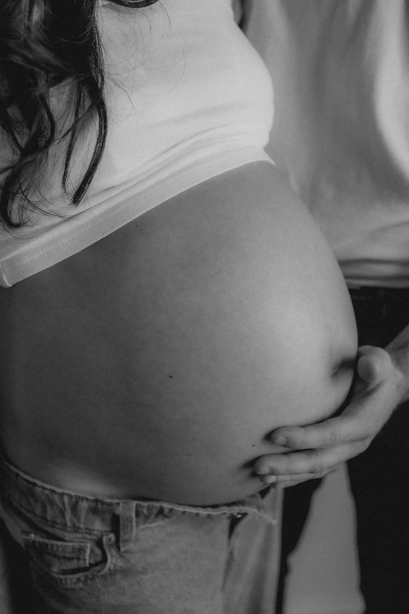 A black and white close-up photo of a pregnant woman’s belly, with one hand resting on her abdomen.