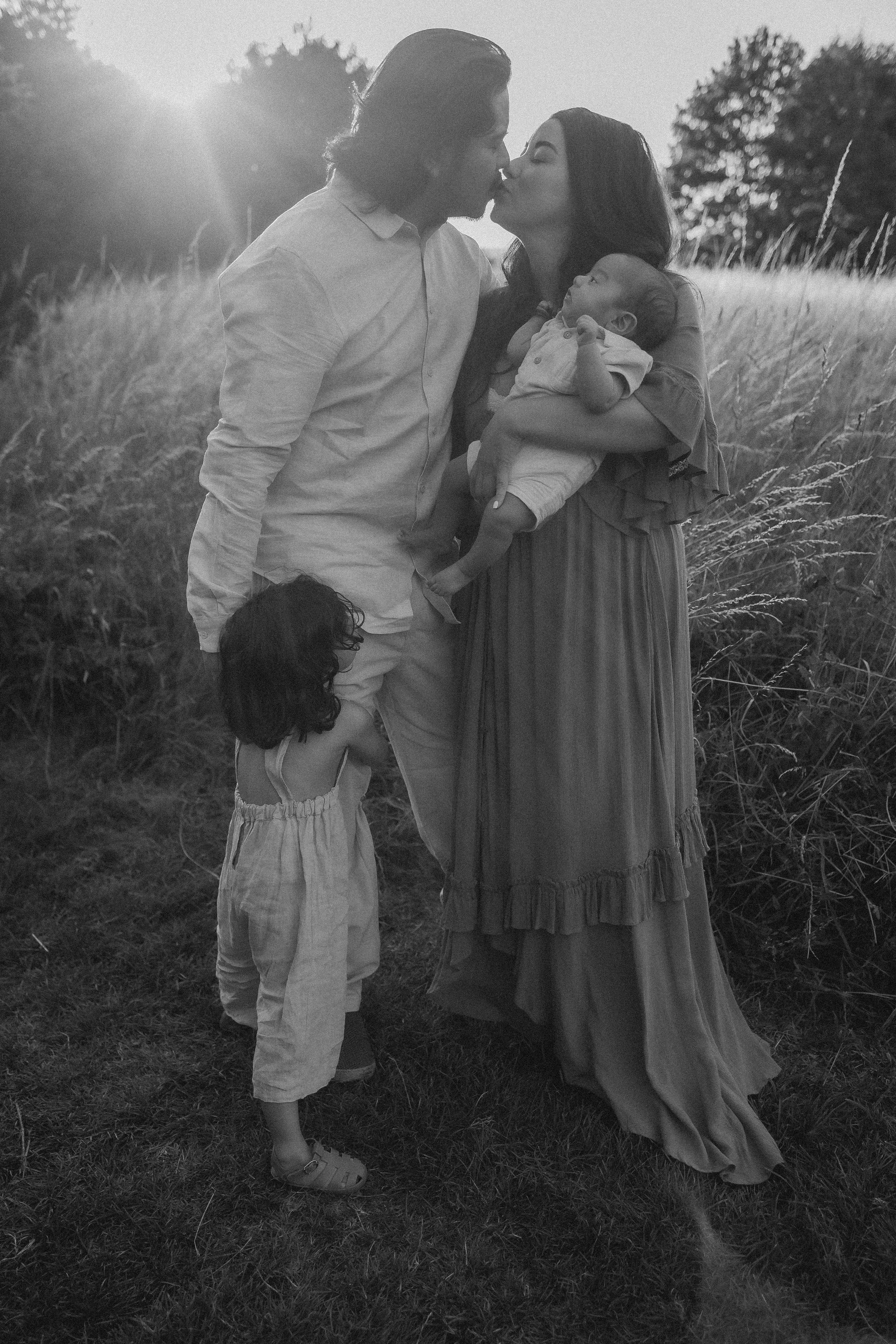A black and white photo of a family outdoors, with a man and woman kissing, a baby in the woman's arms, and a young girl standing at the man's side, set against a field and trees in the background.