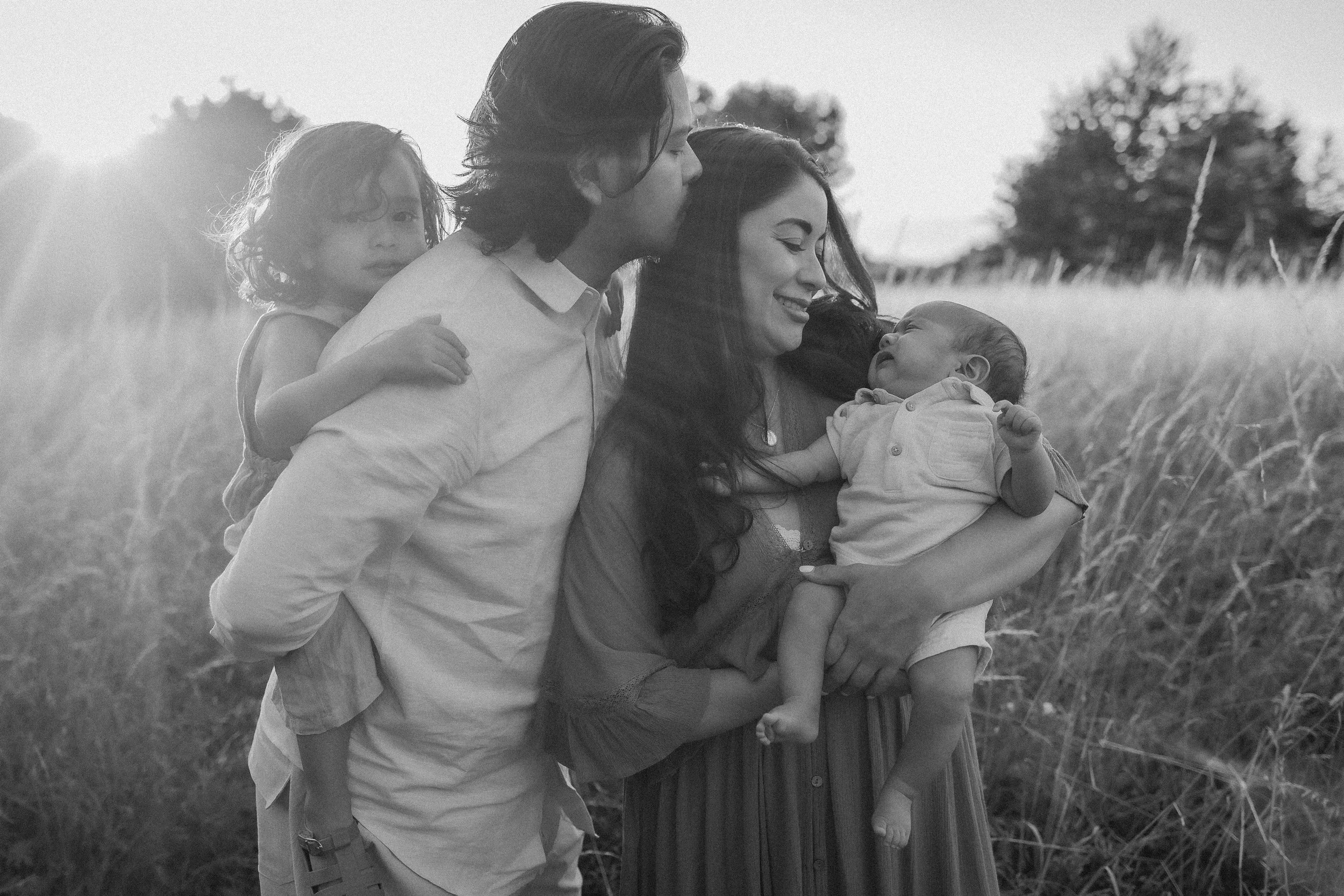 A family of four standing in a field during sunset. The father is kissing the mother's forehead and the mother is holding a baby. A young girl is on the father's back, looking at the camera.