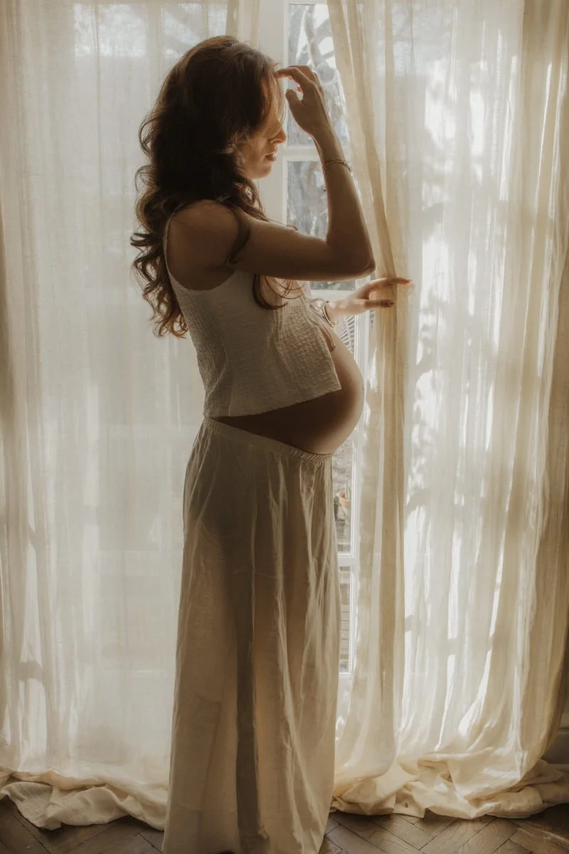 A pregnant woman standing by a window with sheer curtains, looking down and touching her head, wearing a sleeveless top and loose pants.