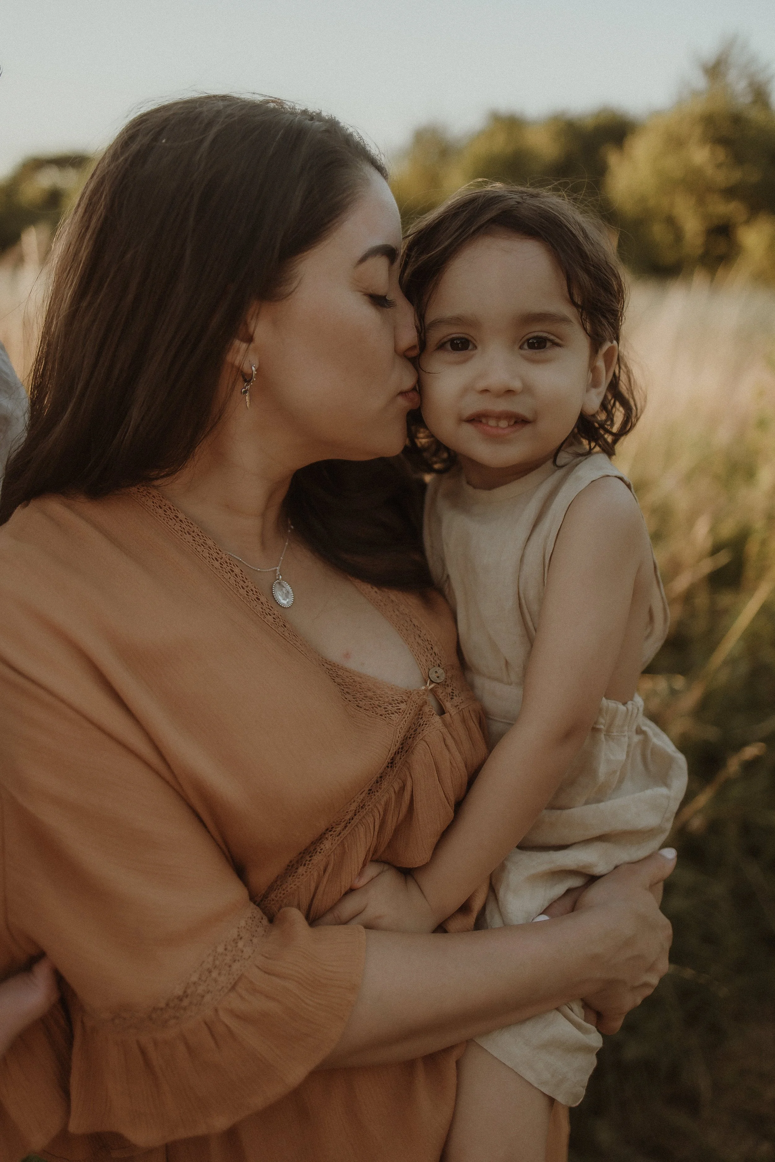 A woman with long dark hair and earrings is kissing a young girl on the cheek outdoors during sunset. The girl has curly hair and is smiling, wearing a beige sleeveless outfit. They are surrounded by tall grass and trees.