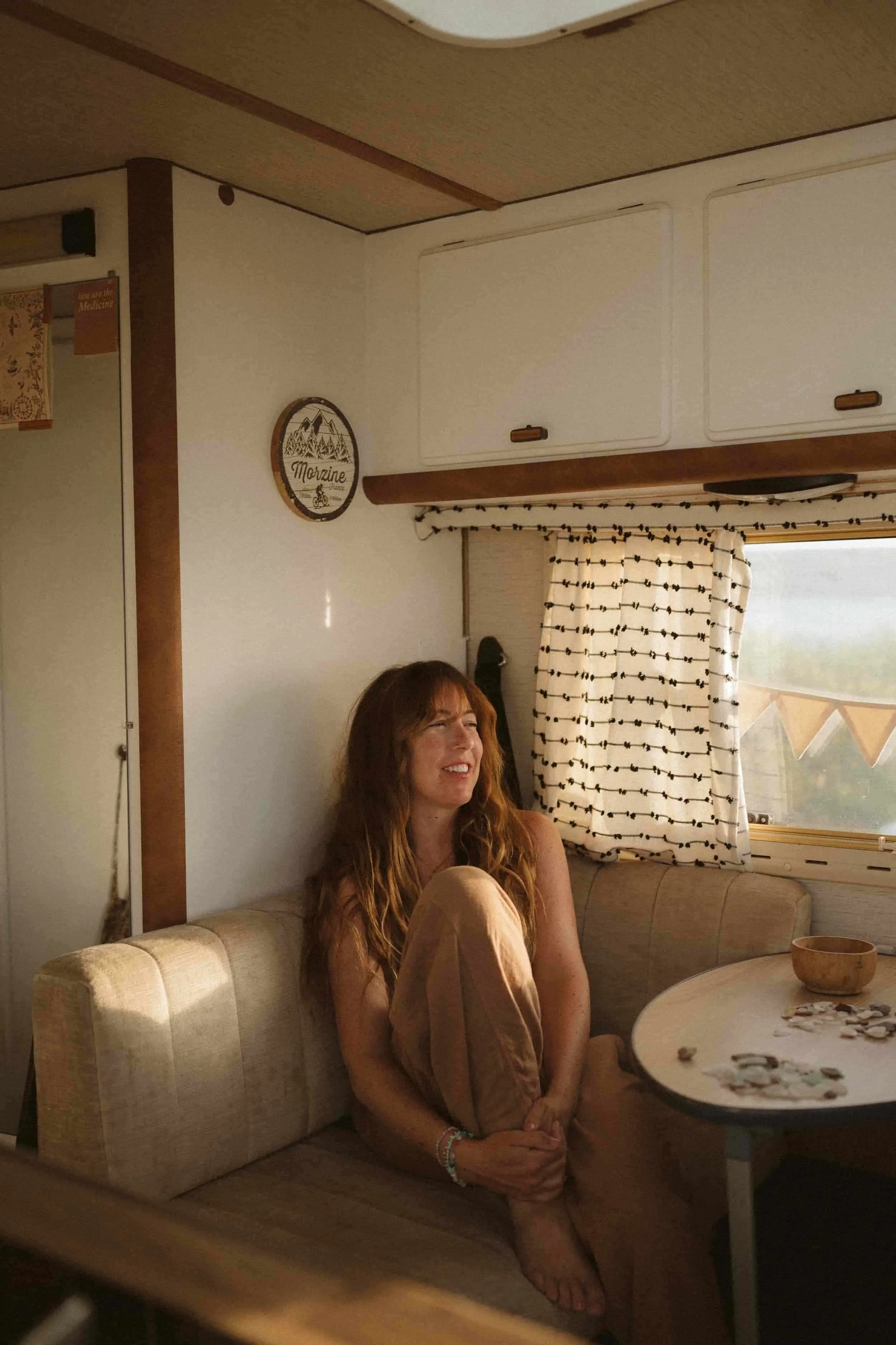 A woman with long red hair sitting on a beige cushioned booth in a room with beige walls, white cabinets, and a window with black and white patterned curtains. She is smiling and looking to her right, with her arms crossed over her stomach and one foot on the floor. A round table in front of her has a wooden bowl and some scattered objects.