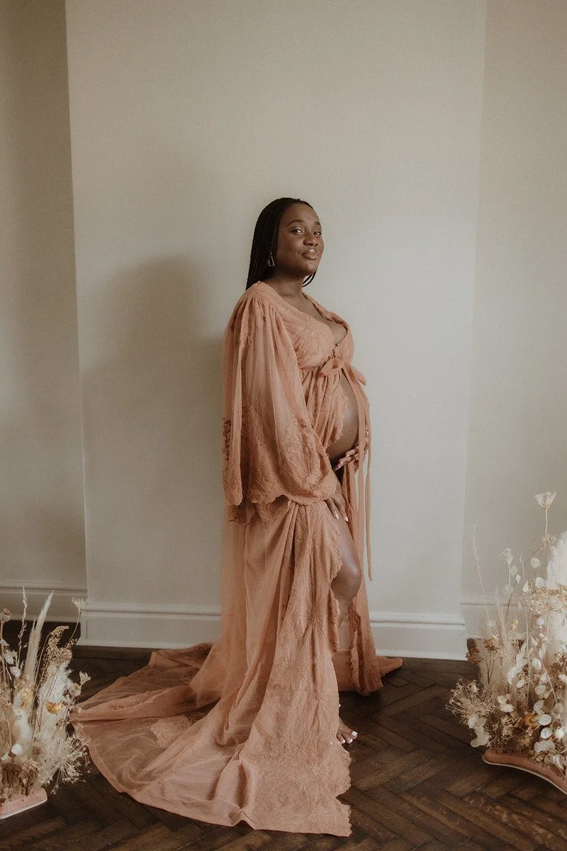 Pregnant woman in a peach-colored dress standing indoors on dark wooden floor, with dried floral arrangements on either side.