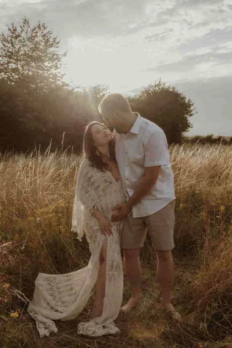 A couple standing in a field during sunset, with the man gently kissing the woman's forehead. The woman is wearing a long, lace dress, and the man is dressed in a white shirt and beige shorts. Trees are in the background under a partly cloudy sky.