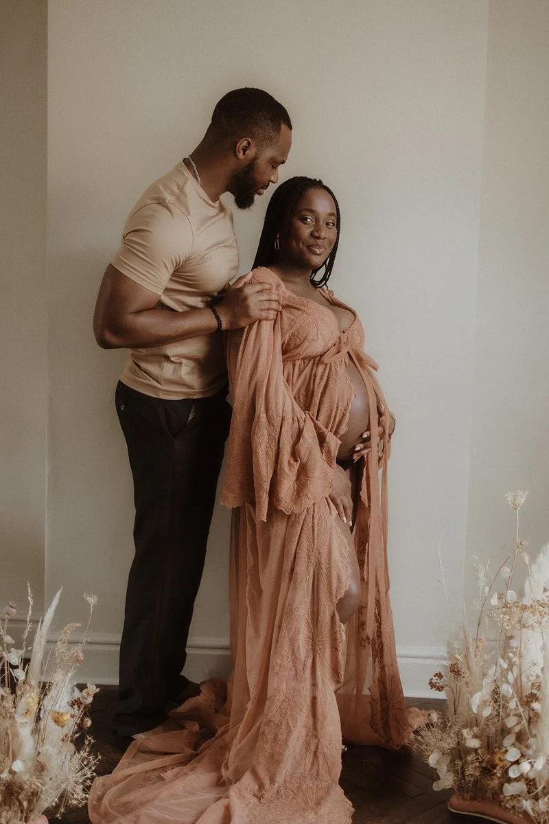 A pregnant woman dressed in a long, flowing peach-colored gown stands next to a man in a beige t-shirt and black pants. The man gently holds the woman’s shoulder, and she is smiling while cradling her belly with both hands. They are standing inside a