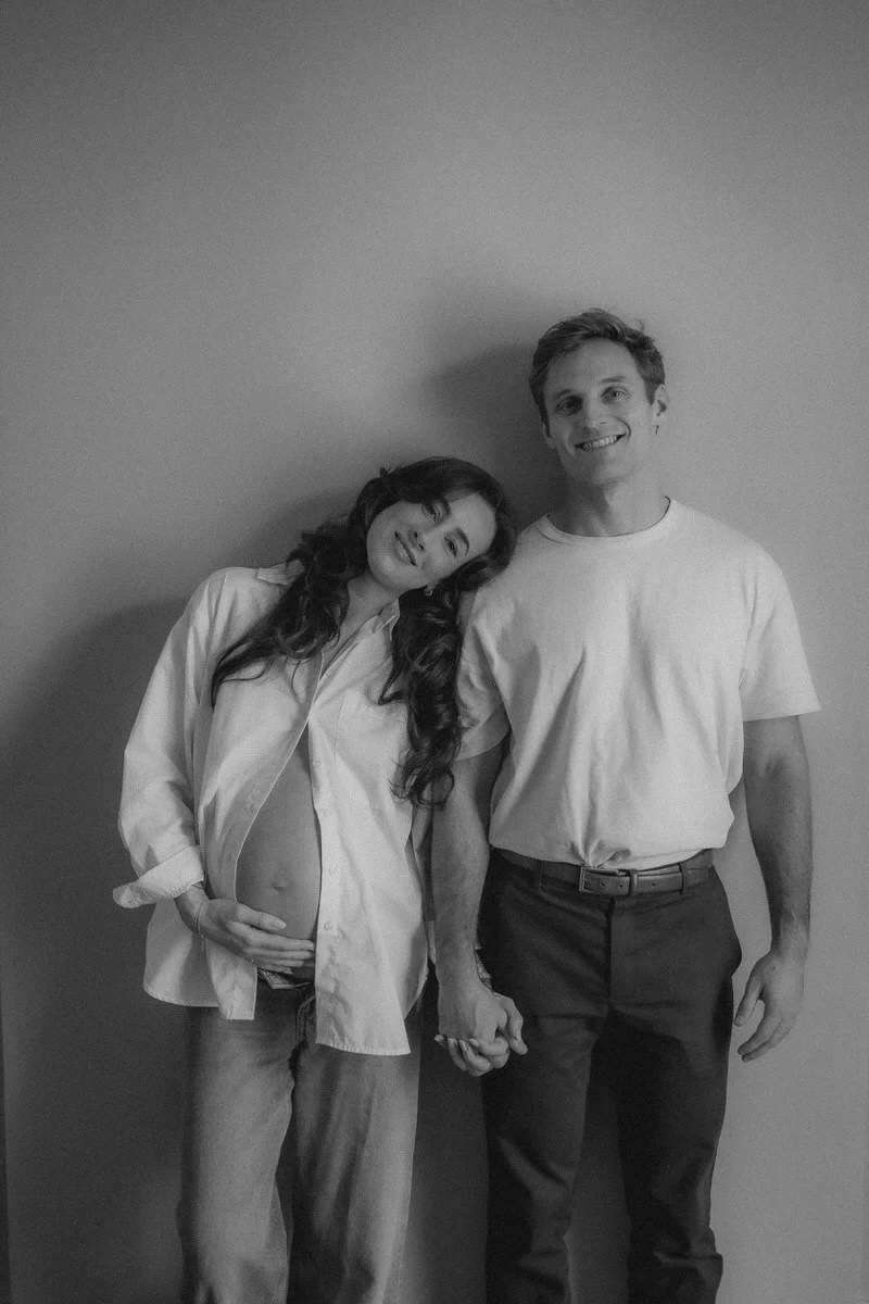 A black and white photo of a smiling couple holding hands, the woman is pregnant and leaning her head on the man's shoulder, standing against a plain wall.