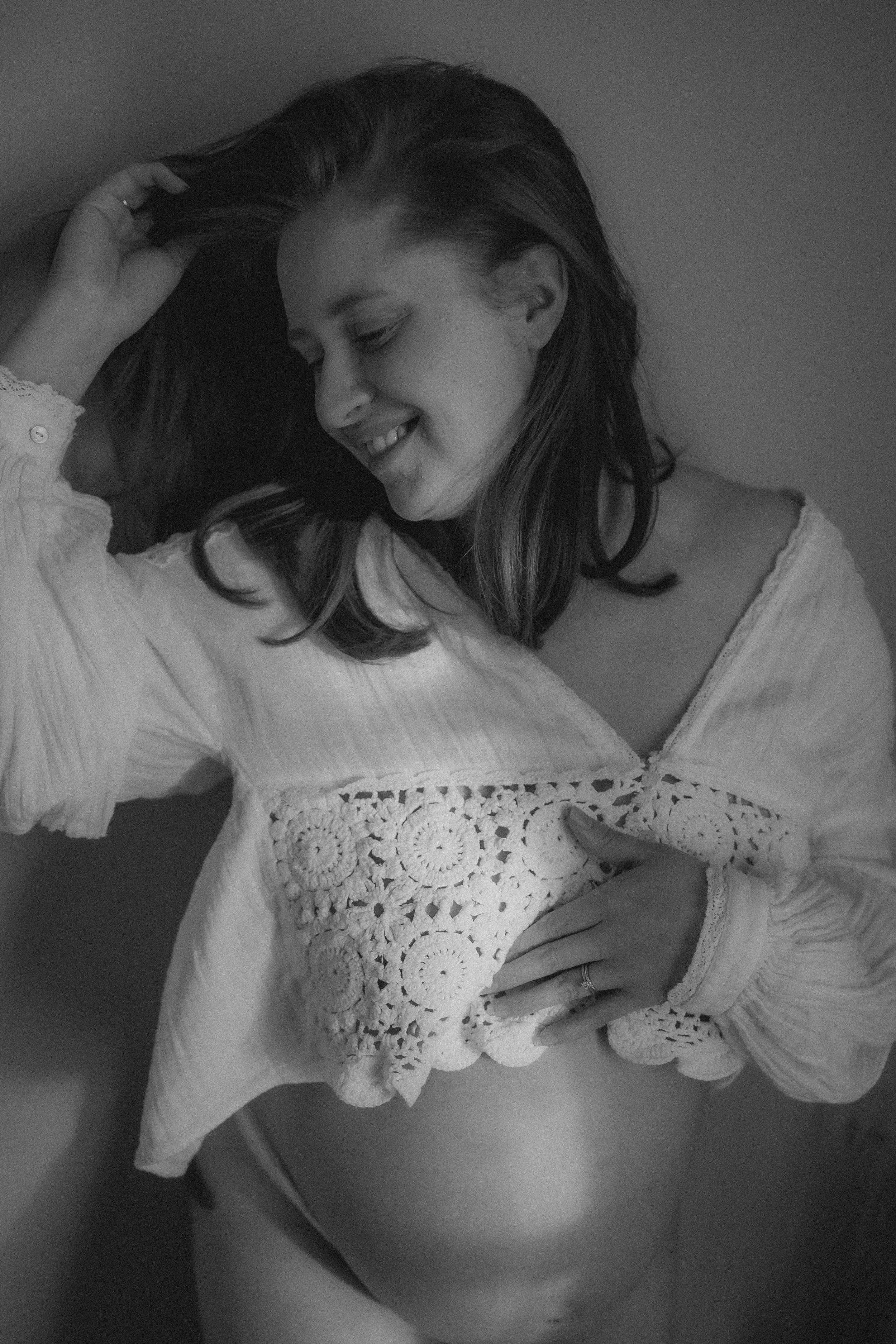 A woman with long hair wearing a crochet and lace top, smiling and hugging herself.