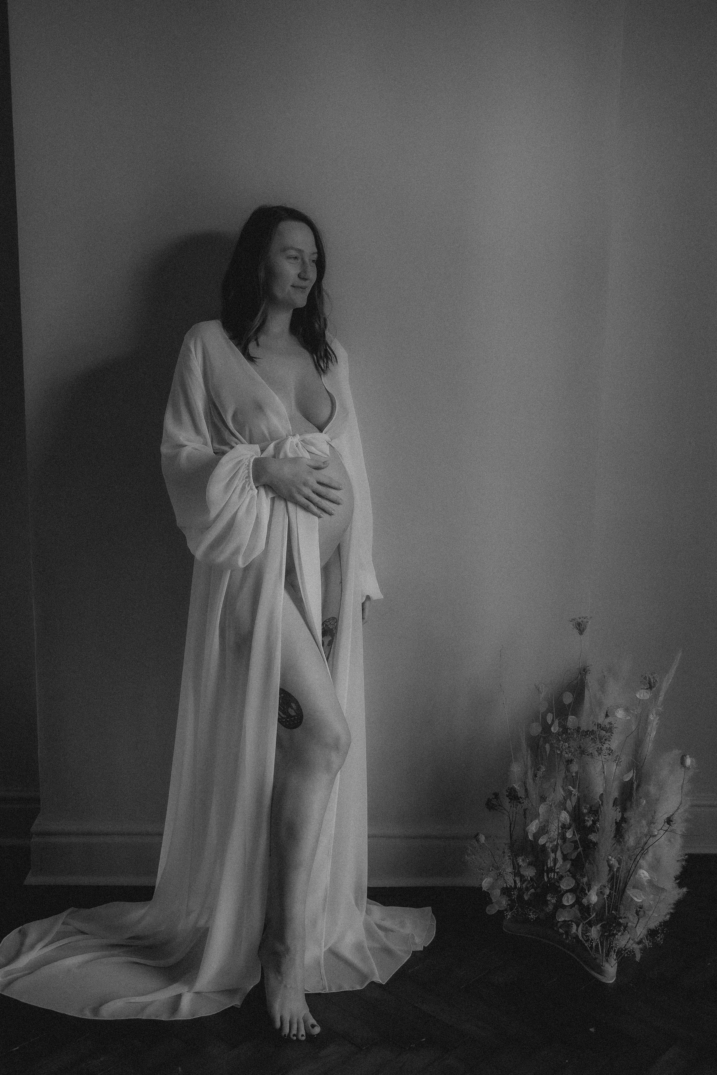 A woman in a long robe stands barefoot next to a vase with flowers against a plain wall.