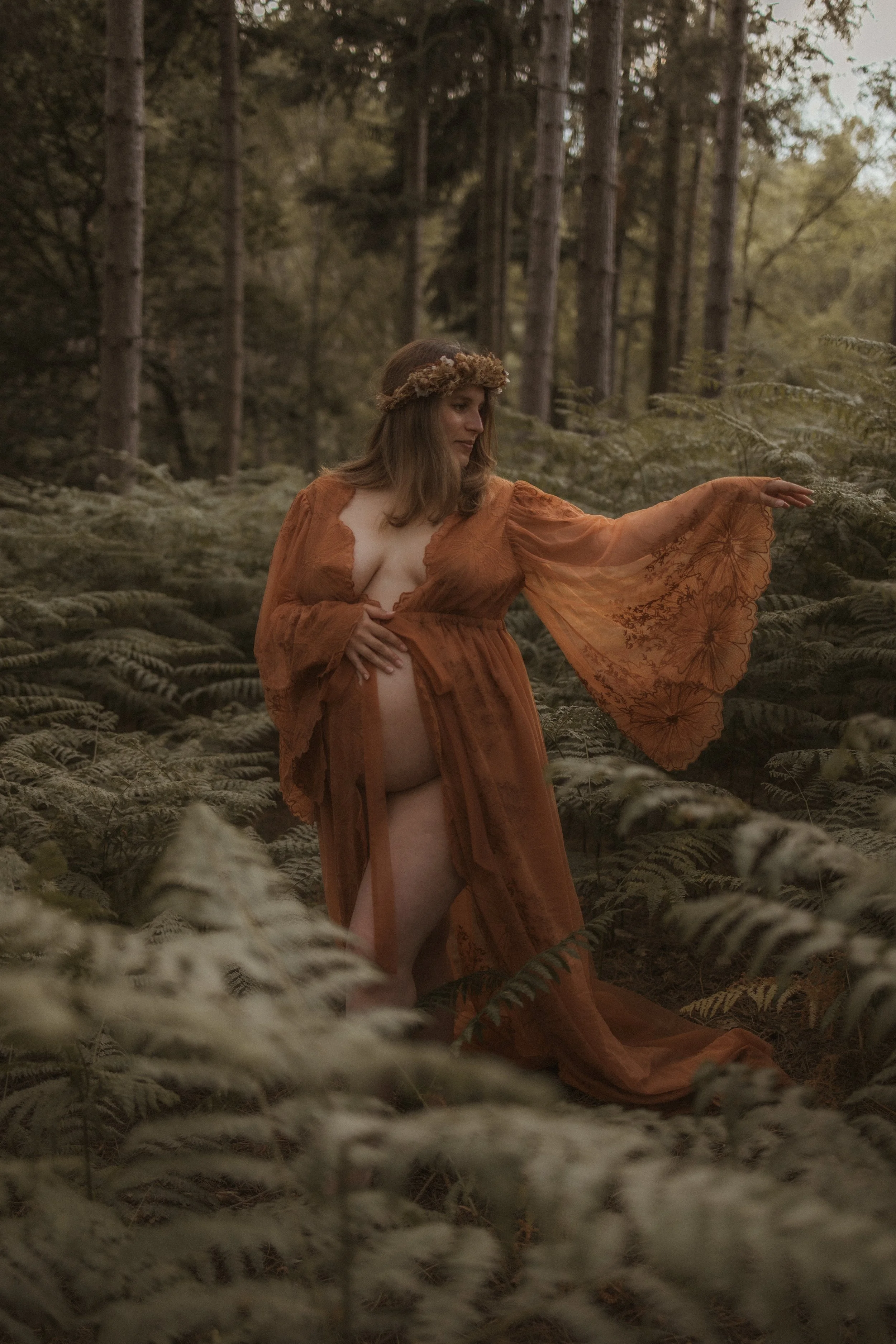 A pregnant woman standing among ferns in a forest, wearing a flower crown and touching nearby foliage.