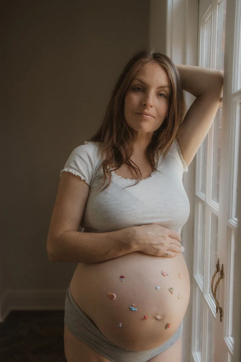 A pregnant woman with brown hair standing in front of a window, gently touching her belly which is decorated with colorful small toys.