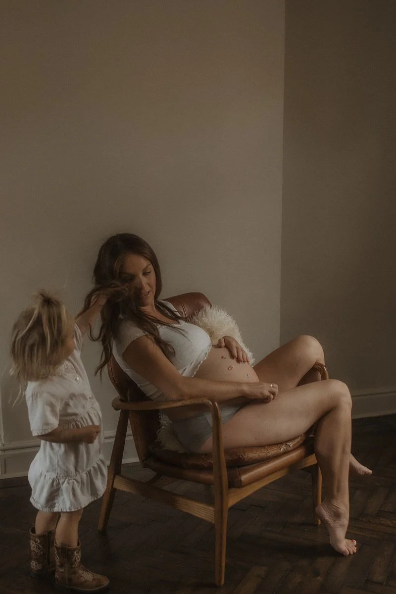 A pregnant woman sitting on a wooden chair with a small child standing beside her, touching her hair in a dimly lit room.