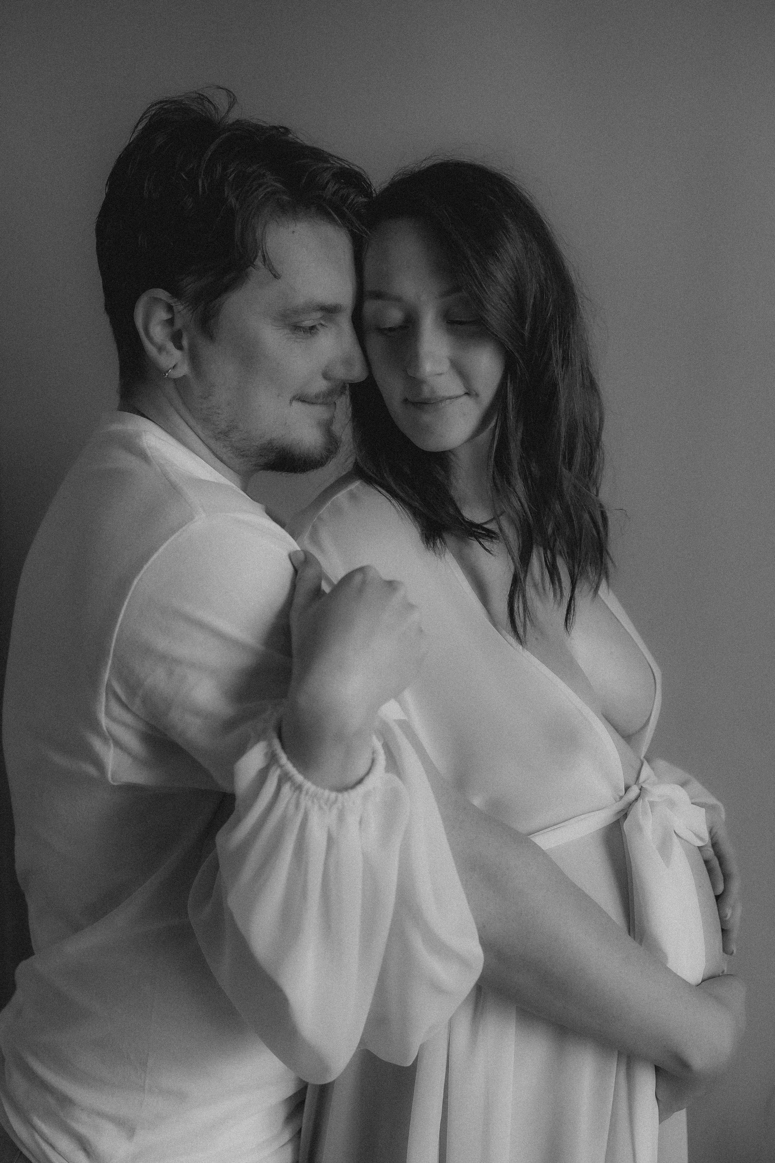 A black and white photo of a couple, with the woman visibly pregnant, embracing and looking lovingly at each other.