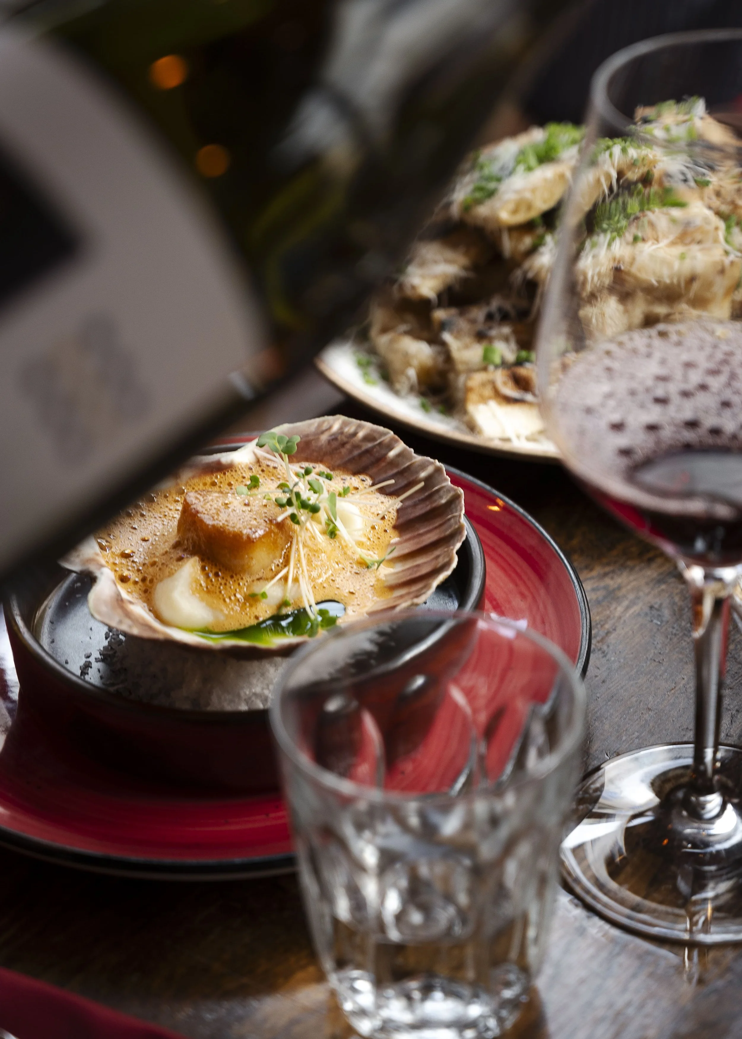 Et serveringsfat med kamskjell, smør, og små krydderurter, omgitt av glas med rødvin.