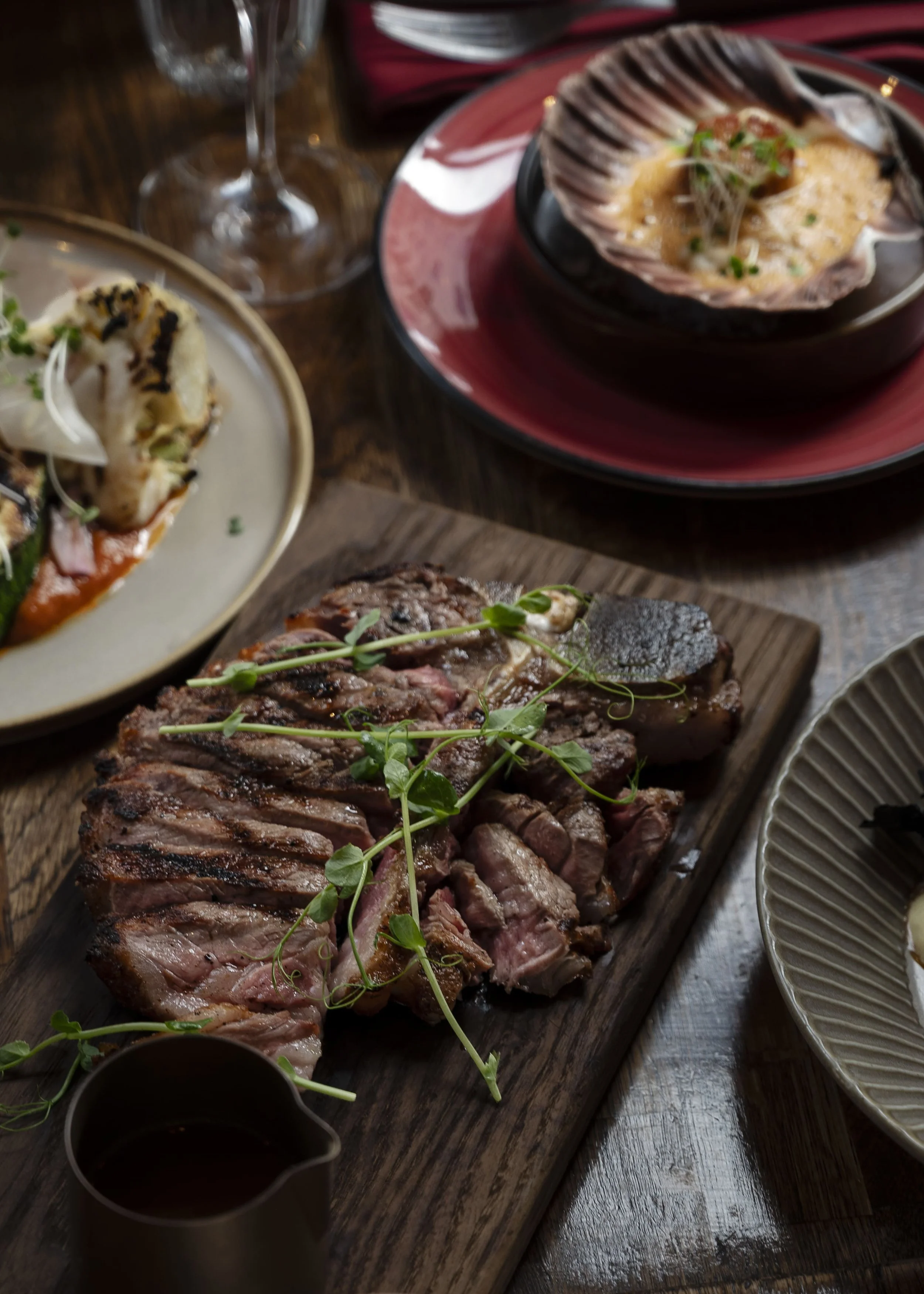 Sliced steak on a wooden board with herbs and butter sauce, scallops au gratin in the shell behind, and small plates at Allmuen Bistro.
