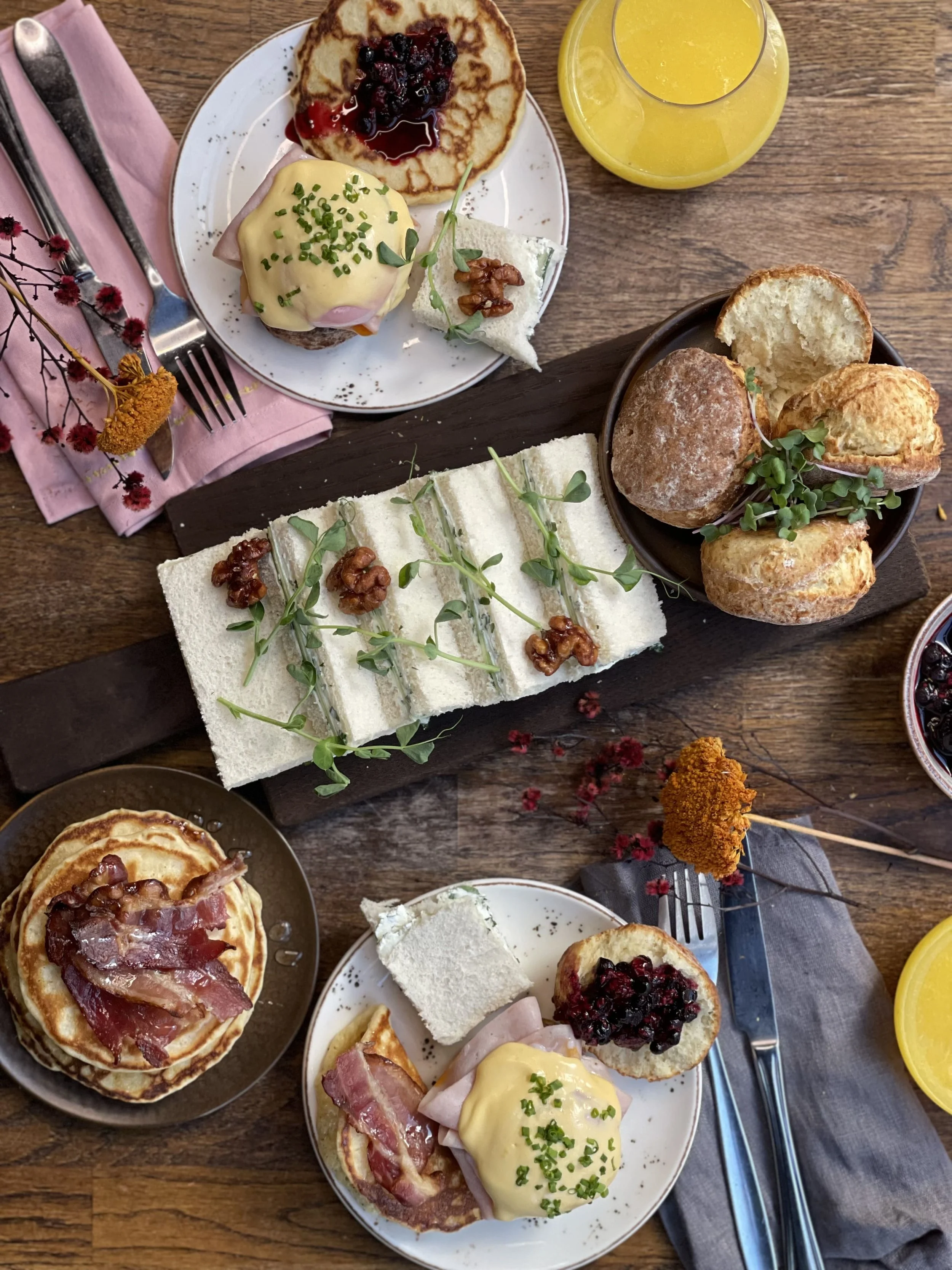 Bilde av lunsjretter tatt ovenfra på et bord. Pannekaker med bacon, eggs benedict, rekke med sandwicher dandert med urter og valnøtter