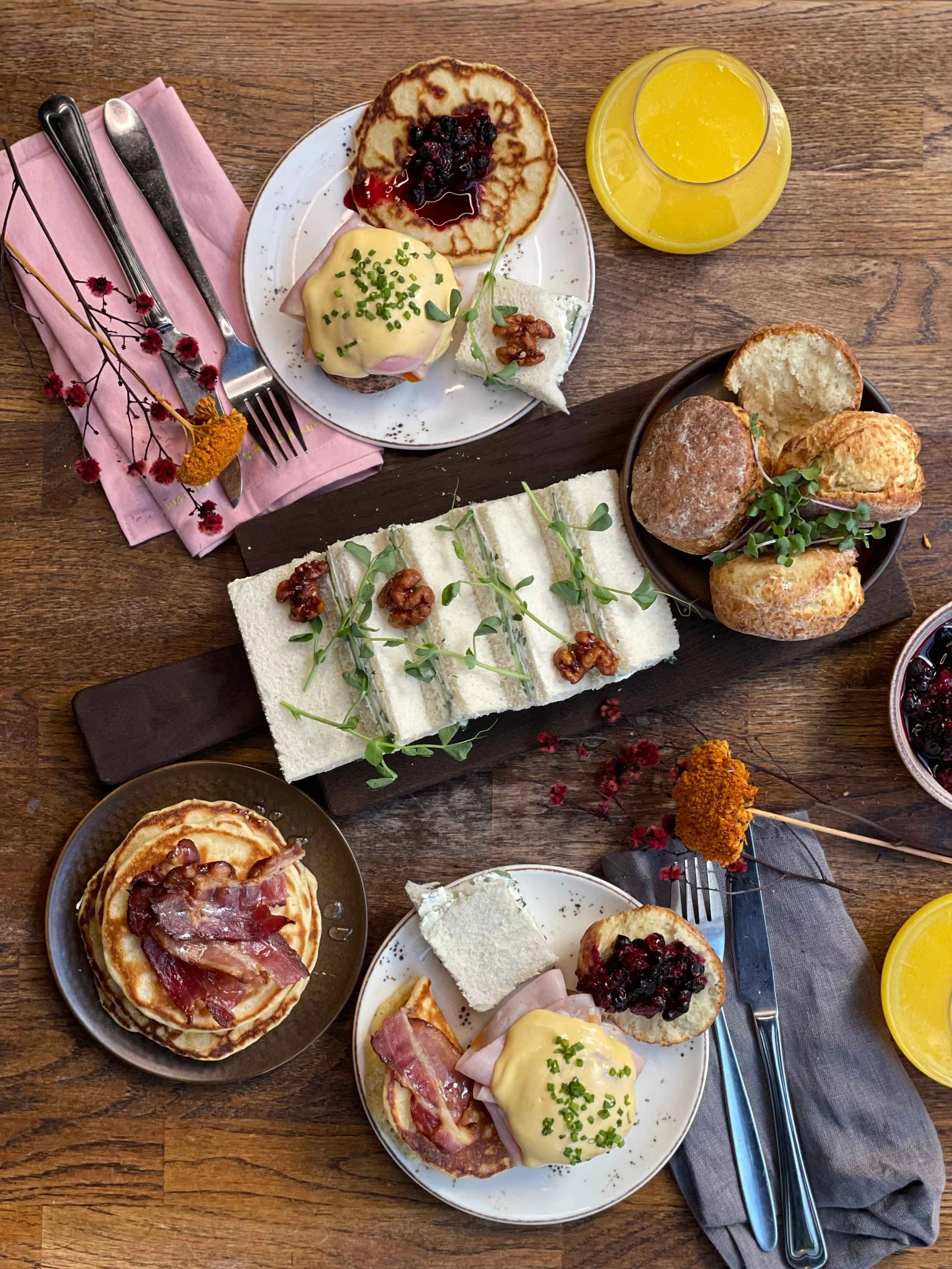 Bilde av lunsjretter tatt ovenfra på et bord. Pannekaker med bacon, eggs benedict, rekke med sandwicher dandert med urter og valnøtter