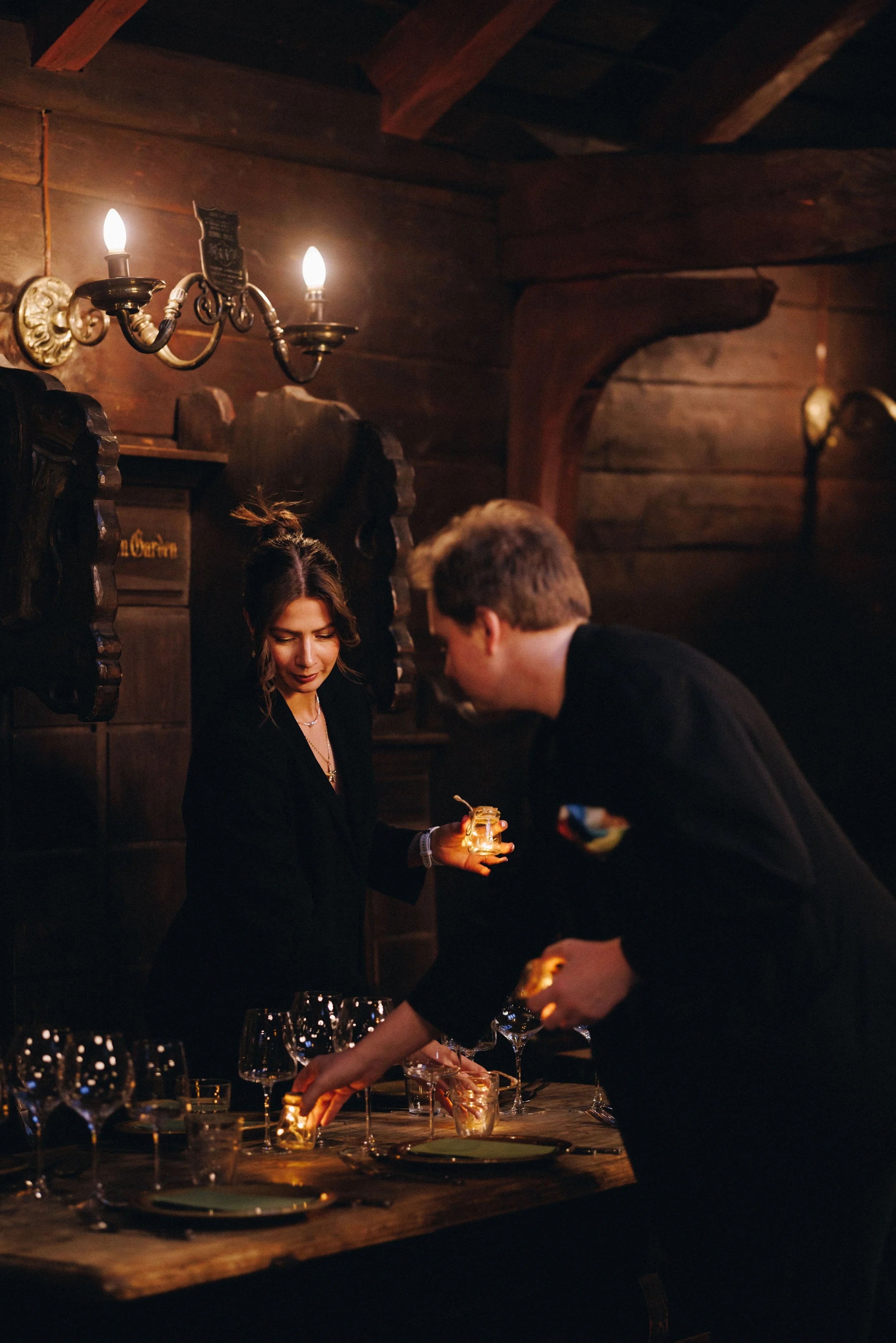 To personer som dekker et bord med vin glasses og lys i en rustikk innendørs setting, muligens i en restaurant eller en privat samling.