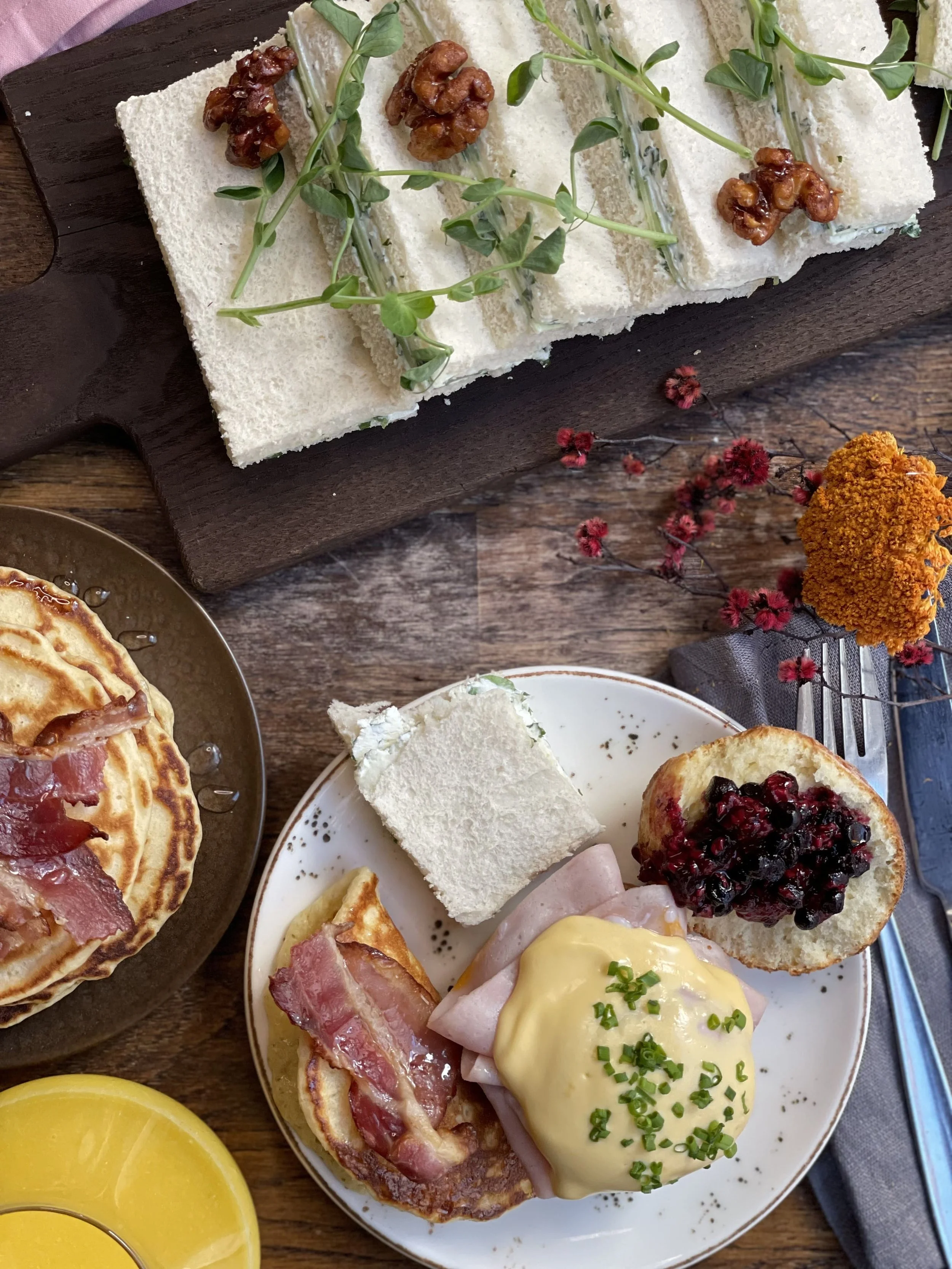Eggs benedict, pannekaker med bacon, rundstykke med syltetøy og sandwich på en tallerken ved siden av et glass juice og dandert fjøl med flere sandwicher.
