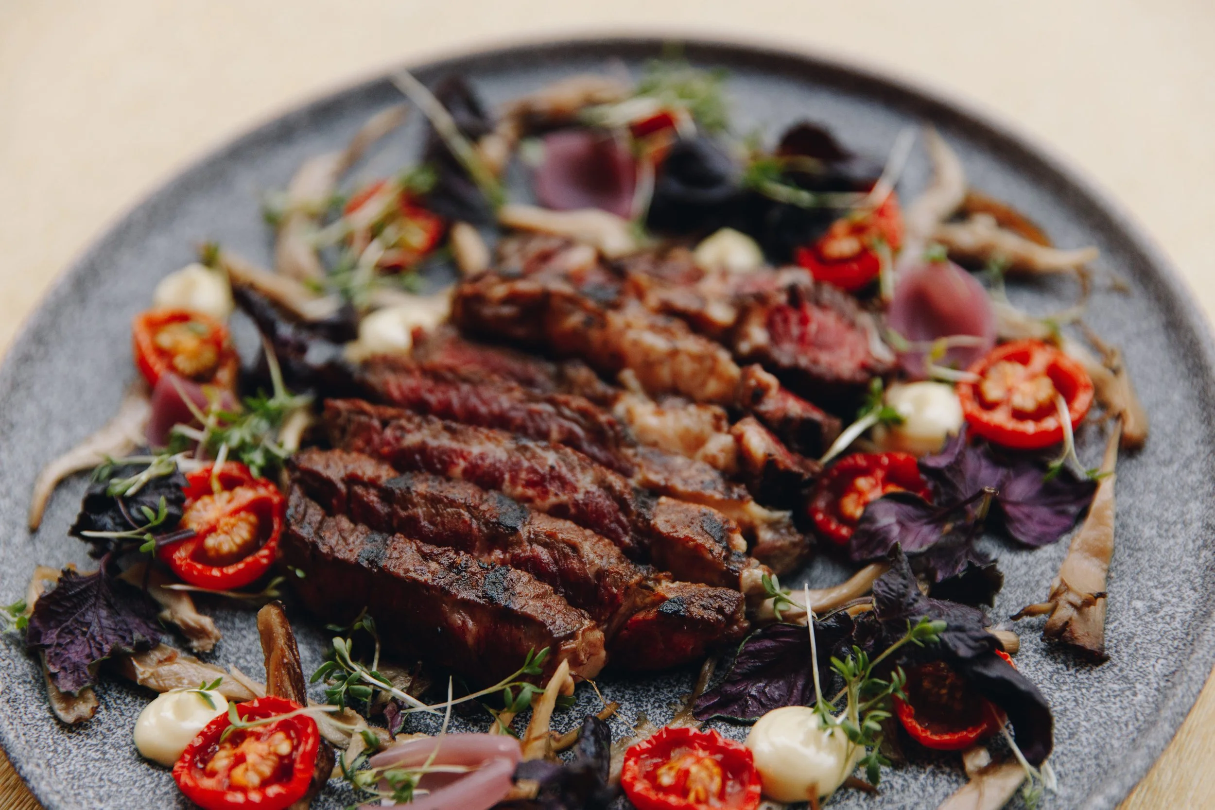 Sliced grilled steak with roasted tomatoes, pickled onions, and fresh herbs on a gray plate, served at Allmuen Bistro.