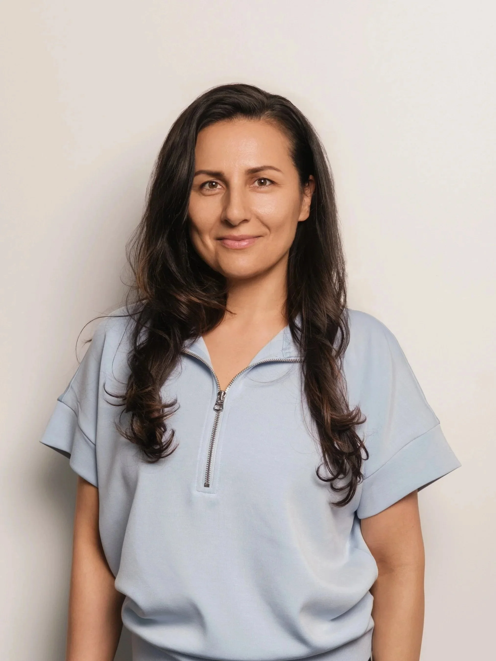 A woman with long dark wavy hair wearing a light blue zip-up top standing against a plain white background.