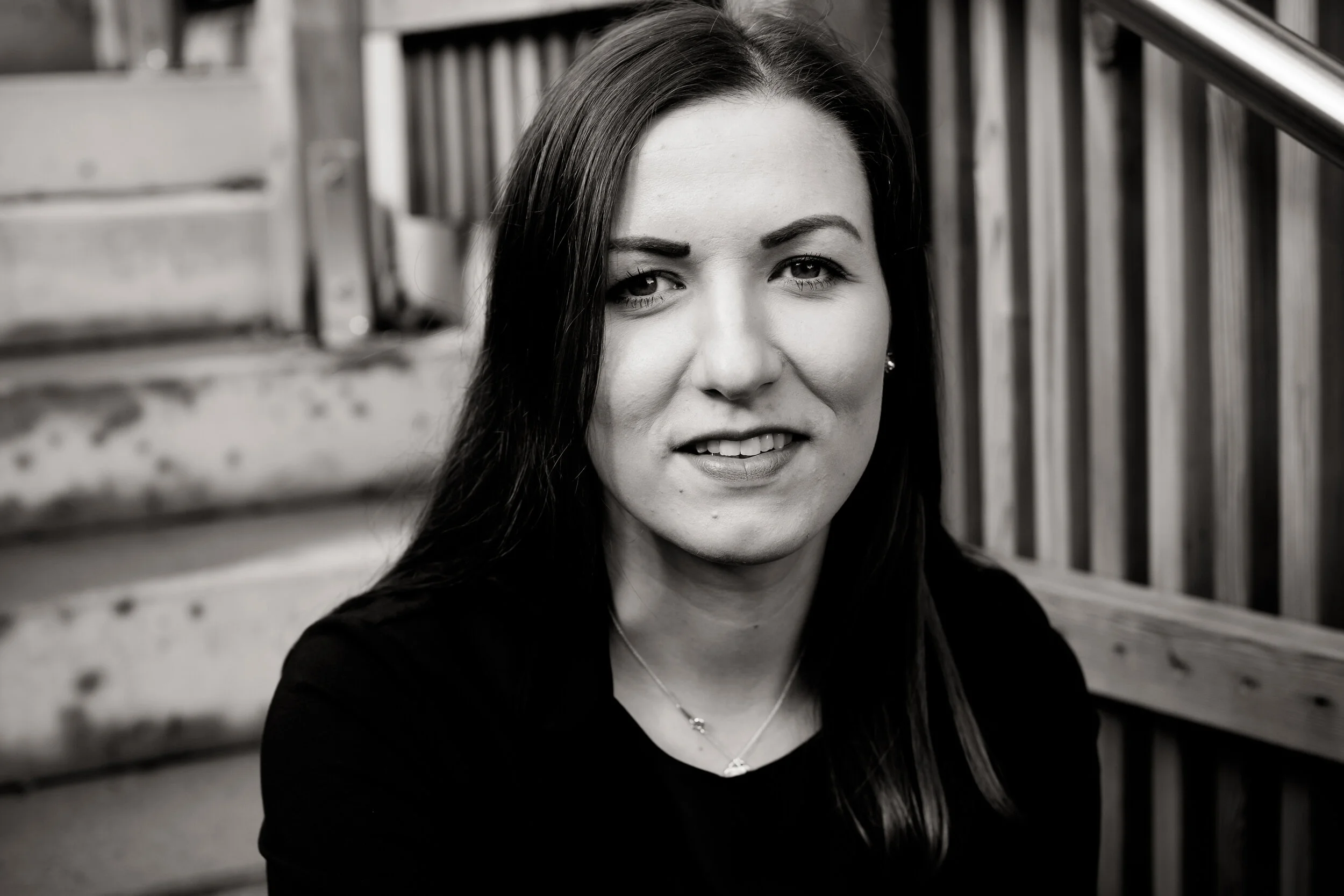 Black and white photo of a woman with dark hair, sitting on outdoor steps, smiling slightly, wearing a dark top and a necklace.
