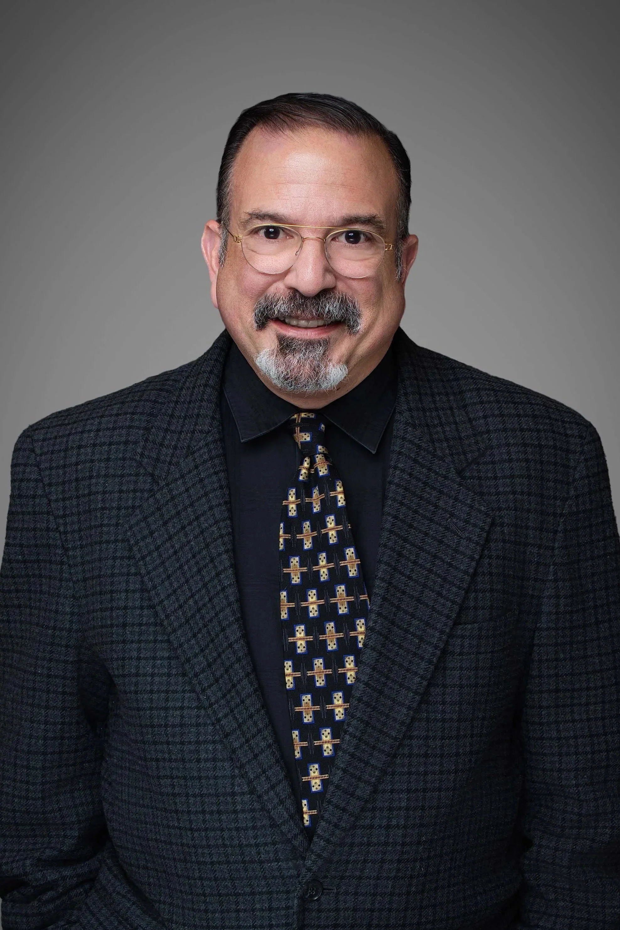 A middle-aged man with dark hair, glasses, and a beard and mustache, smiling at the camera, wearing a patterned blazer, dark shirt, and a tie with geometric designs, against a plain background.