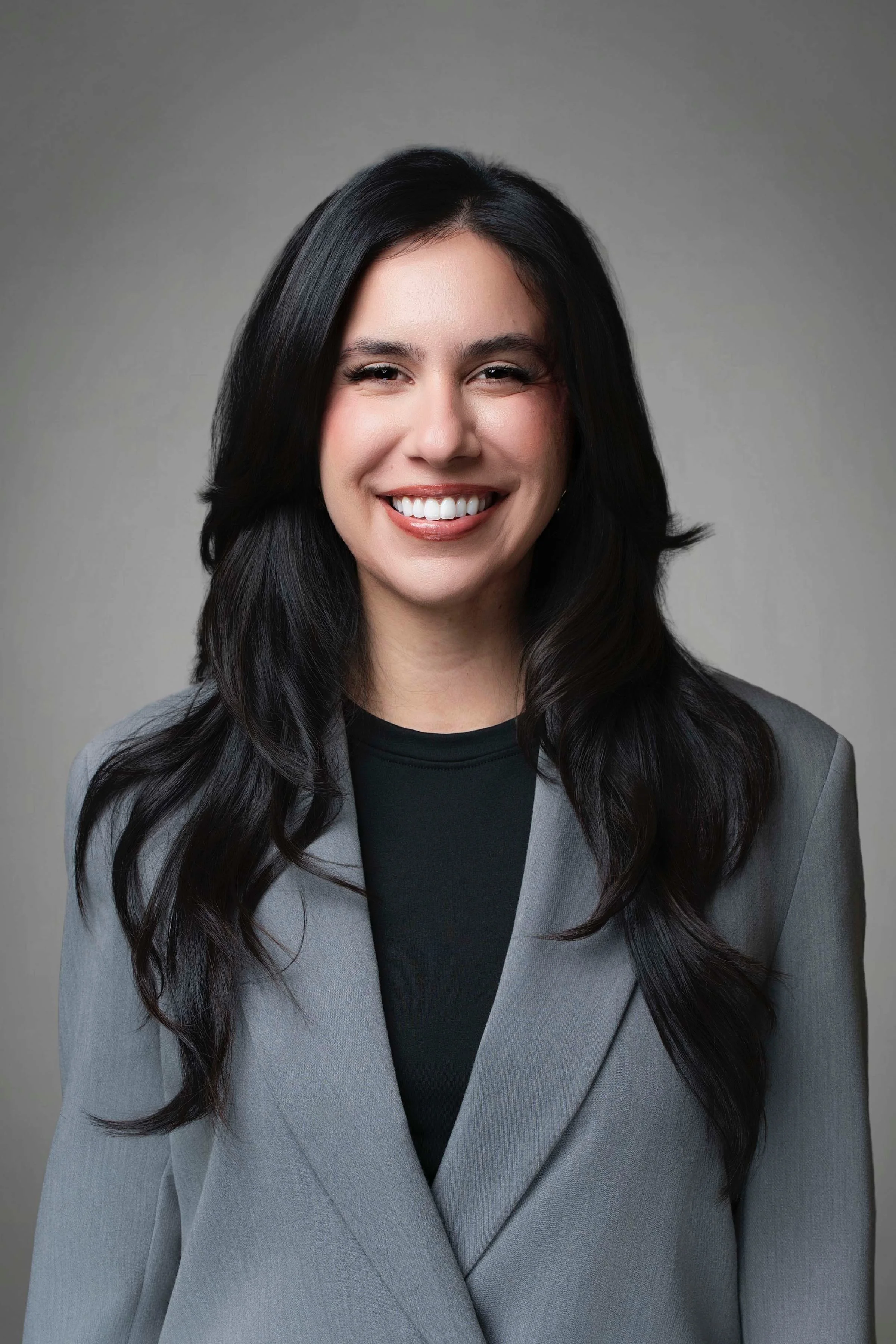 Portrait of a smiling woman with long dark hair wearing a gray blazer and black top against a neutral background.