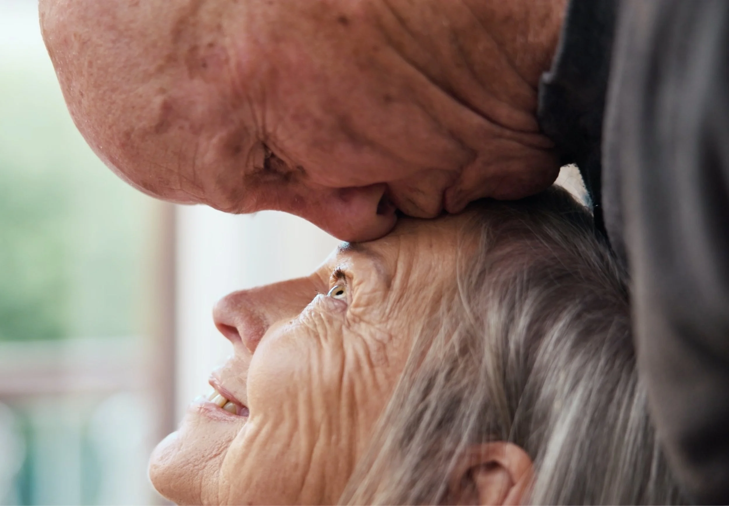 An elderly woman with gray hair smiling as she shares a close, affectionate moment with an older man, who is gently kissing her on the forehead.