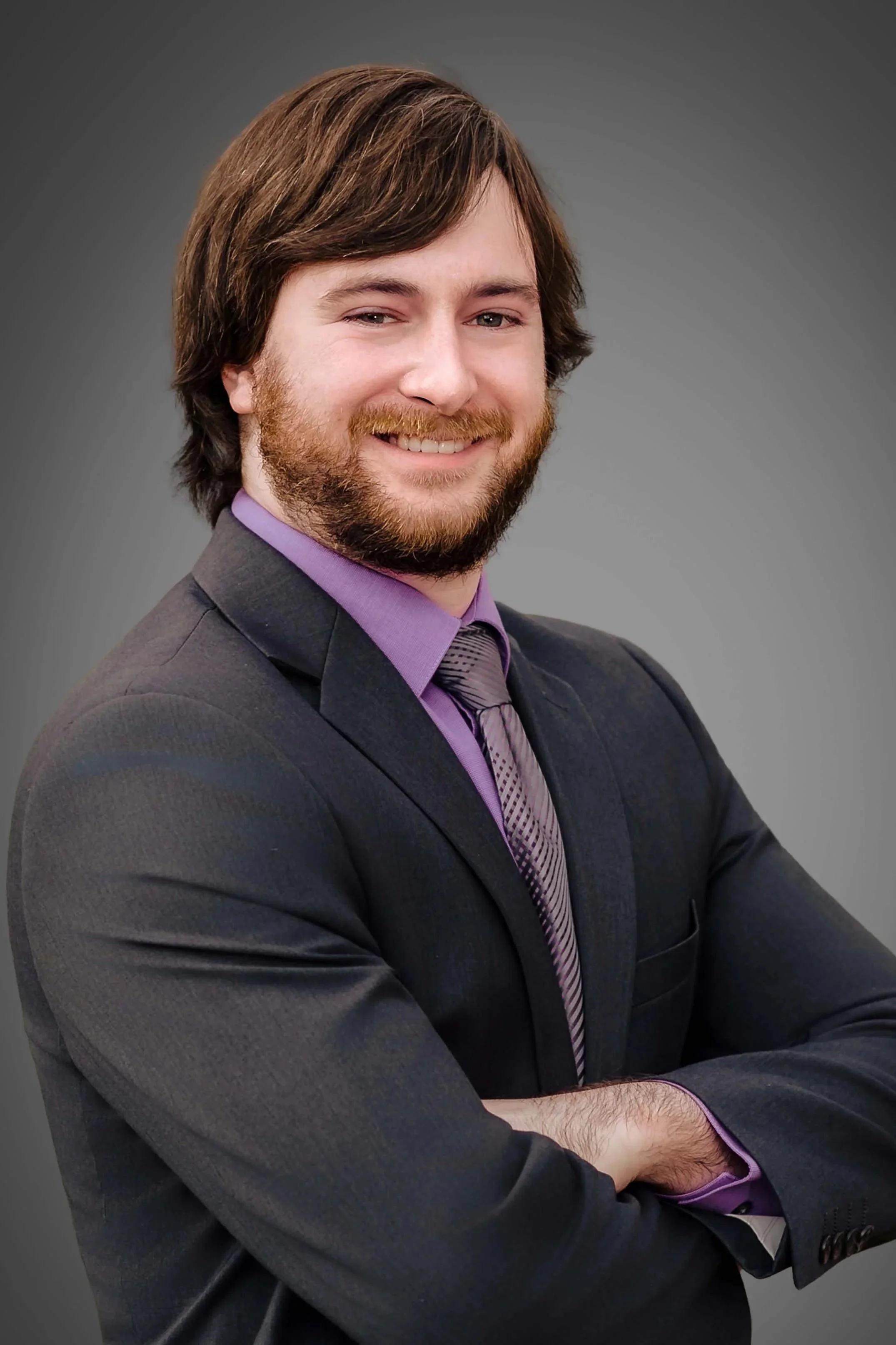A young man with brown hair and a beard, smiling, dressed in a dark suit, purple dress shirt, and patterned purple tie, posing with arms crossed against a gray background.