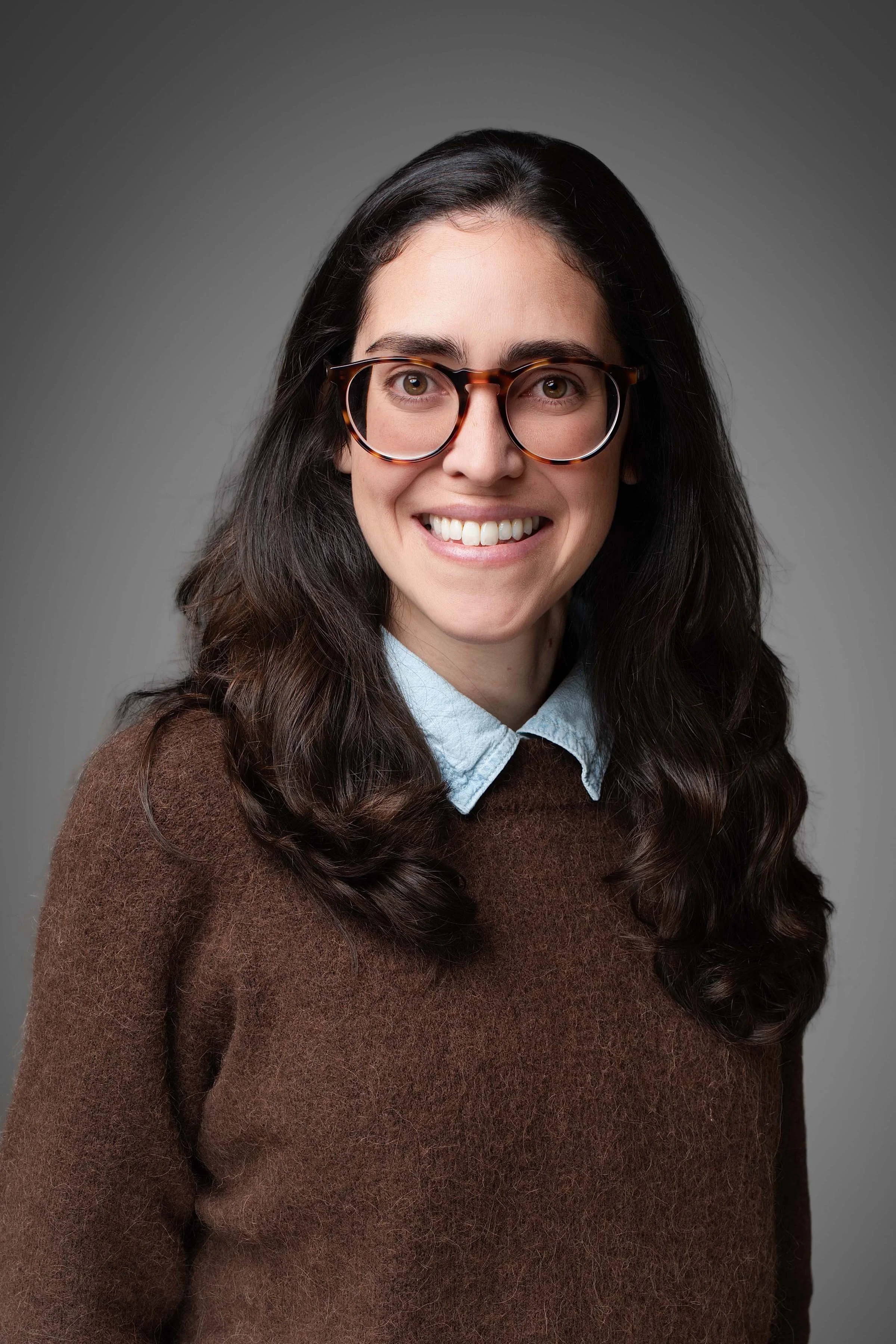 A woman with long dark hair, smiling, wearing a brown sweater and a light blue collared shirt, standing against a gray background.