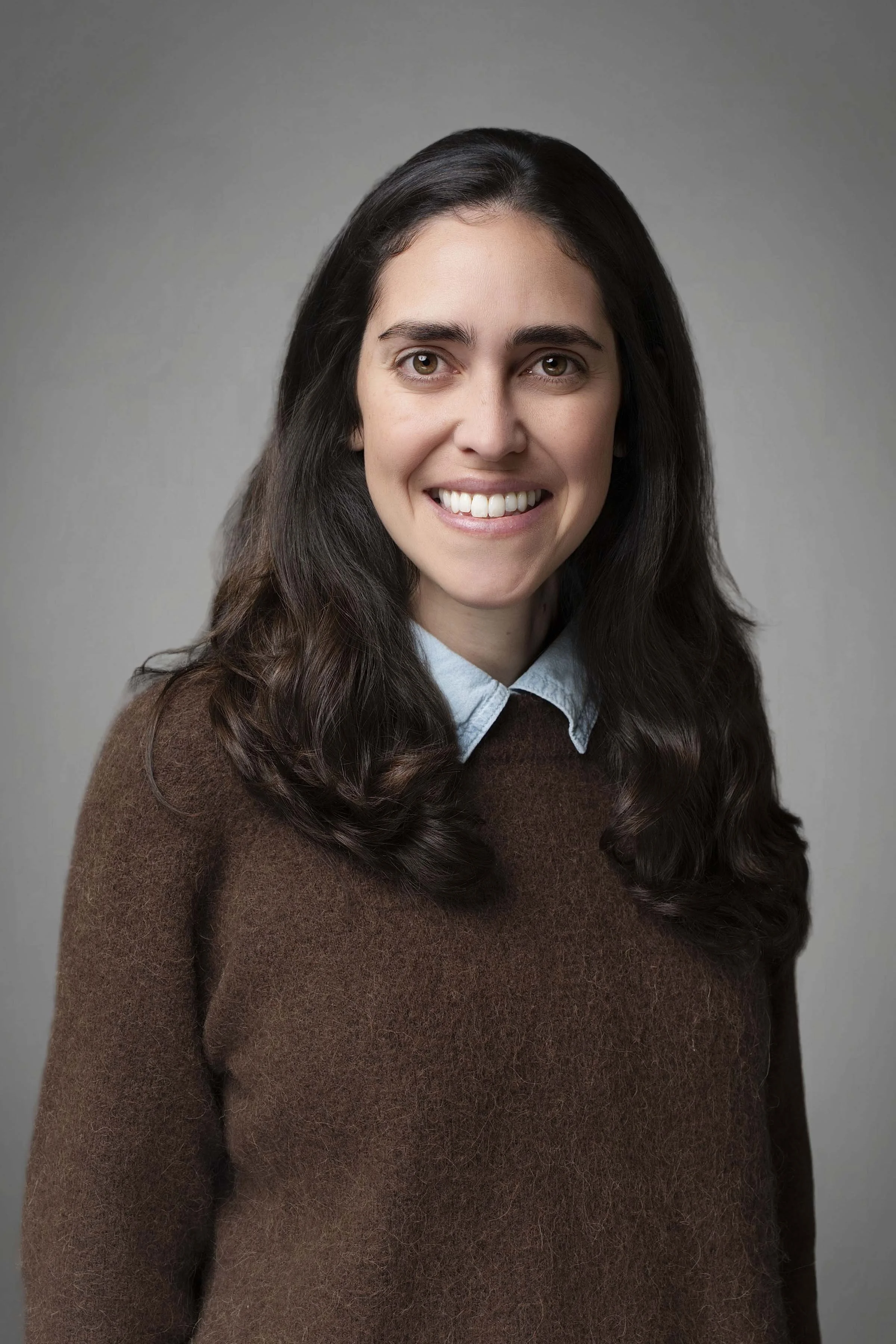 A woman with long dark hair, smiling, wearing a brown sweater and a light blue collared shirt, standing against a gray background.